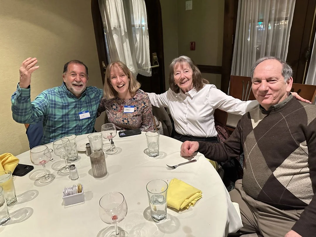 Four adults sitting at a round dining table, smiling and celebrating; the table has wine glasses, water glasses, a yellow napkin, a salt shaker, and utensil. The background shows windows with white curtains.