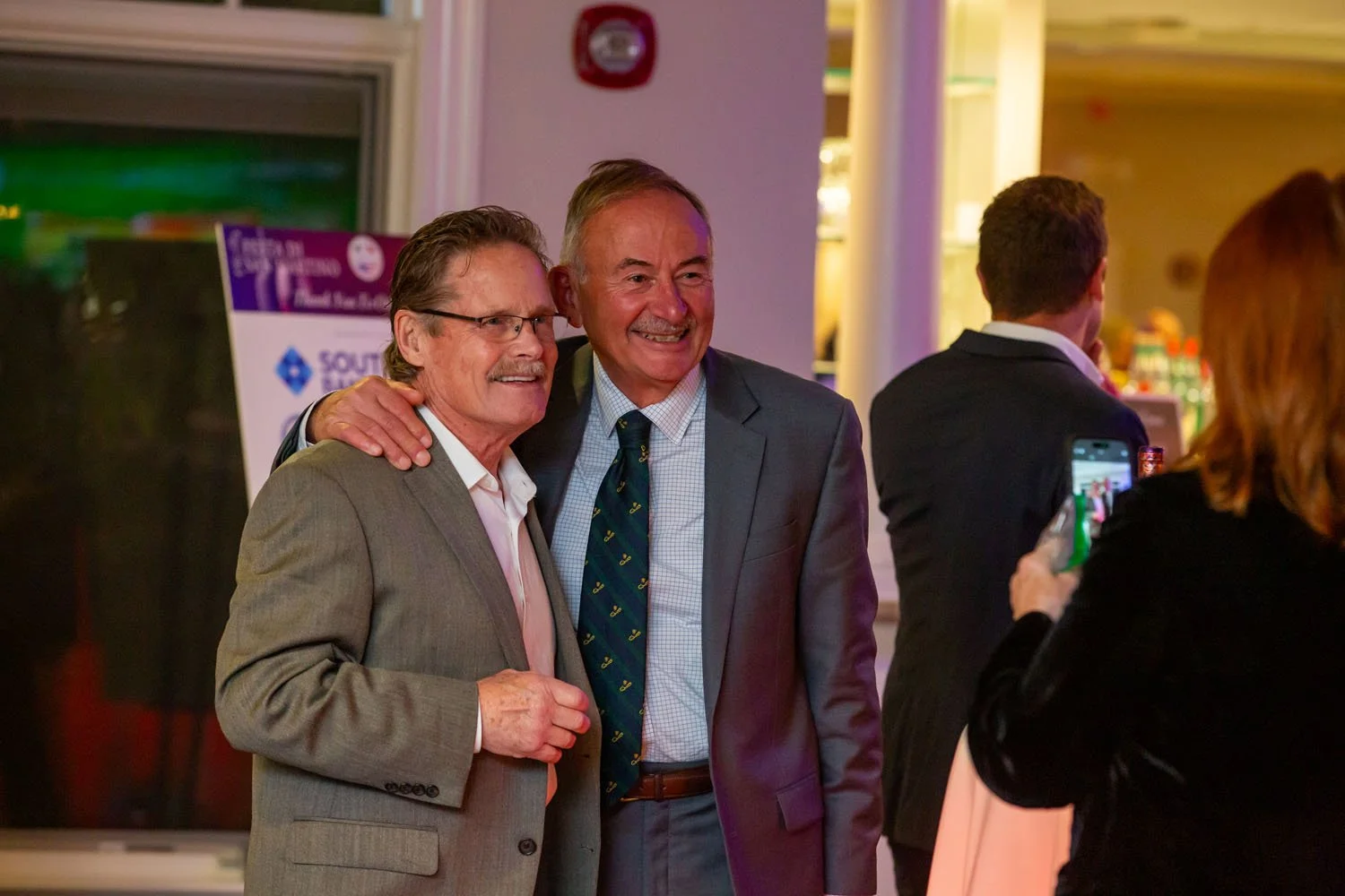 Two men in suits smiling and posing for a photo at an event, with one man having his arm around the other.