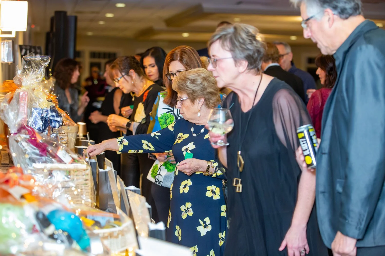 People at a charity auction looking at items on a table, some holding drinks, in an indoor event space.