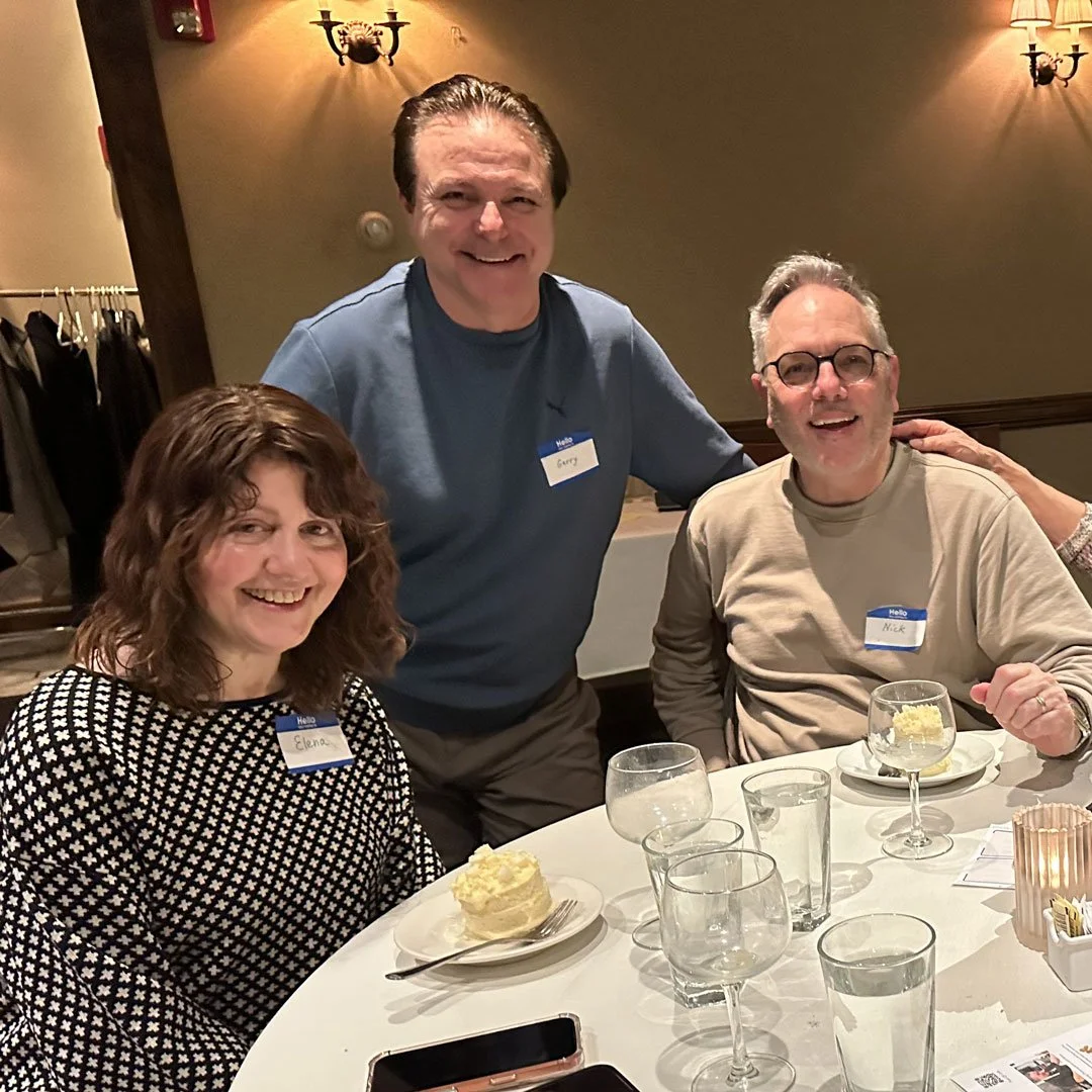 Three people smiling at a table during a social gathering. The woman on the left has curly brown hair and is wearing a black and white patterned top. The man in the middle has dark hair and is wearing a blue shirt. The man on the right has glasses, g
