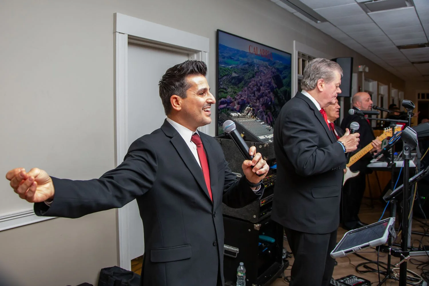 Men in black suits and red ties singing into microphones during a live band performance, with a soundboard, guitar, and large monitor screen in the background.