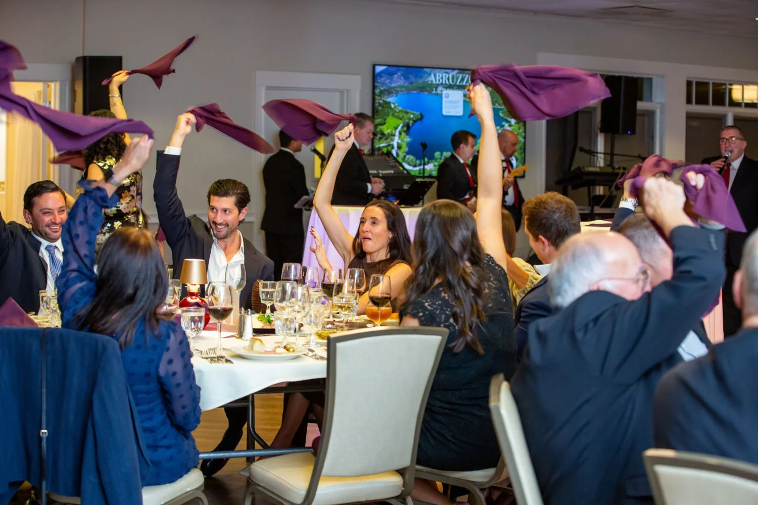 People at a celebration or dinner party are waving purple cloths in the air while seated at tables with drinks and plates, with a band performing live music in the background.