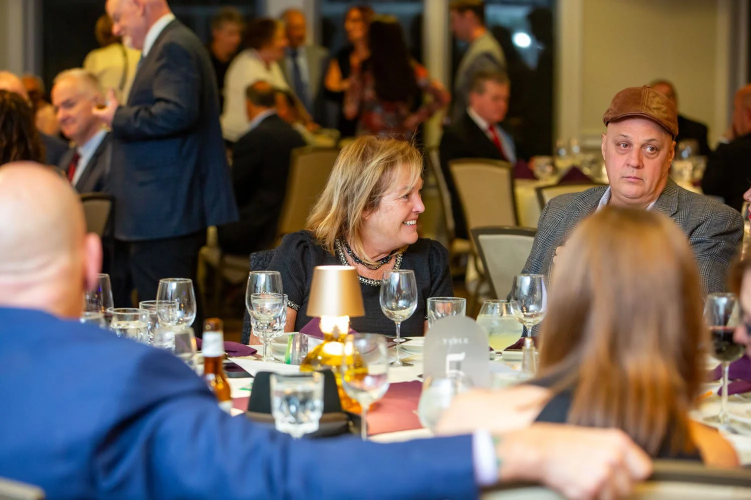 People gathered at a formal event, sitting at tables with glassware and decorative items, engaged in conversation, with some standing and mingling in the background.
