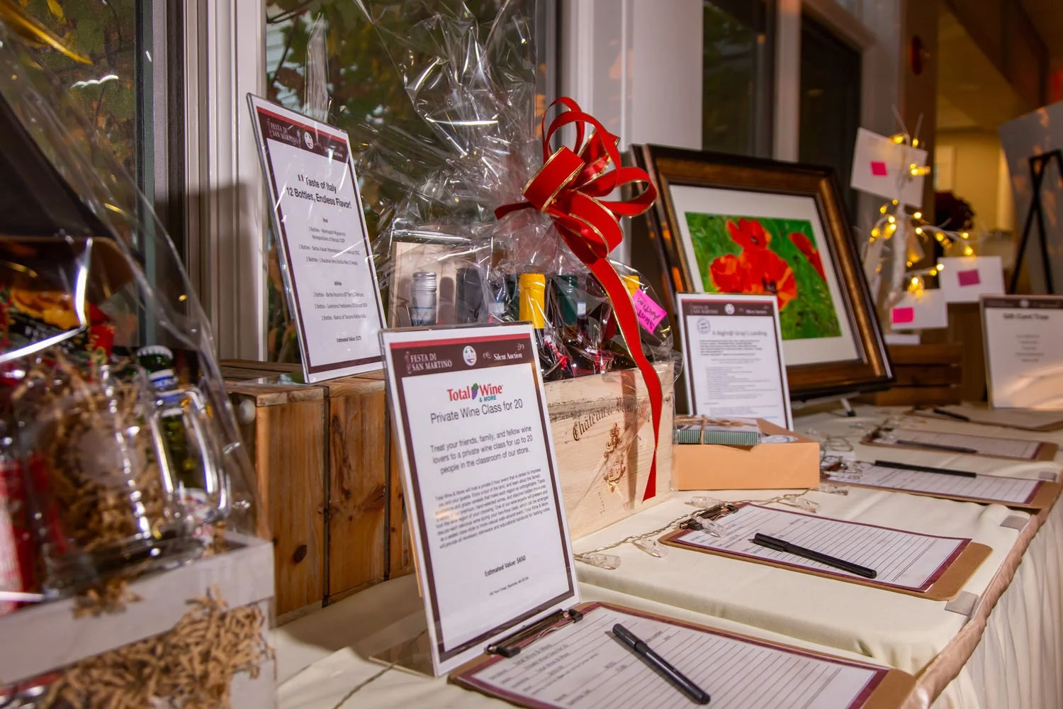 Table with items for a silent auction, including wrapped gift baskets, framed artwork, and informational signs, with clipboards and pens for bidding.