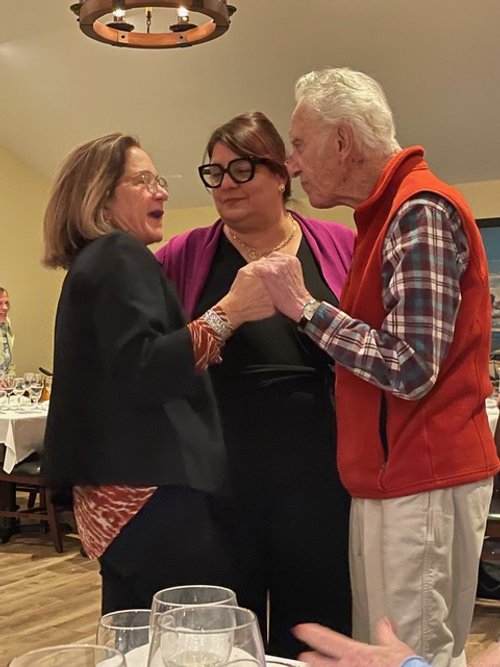 Three people standing closely together, engaged in conversation at a social event. One person, wearing a red vest, is holding the hand of another person in a patterned shirt and dark jacket. A third person in glasses and a black outfit looks on. Ther