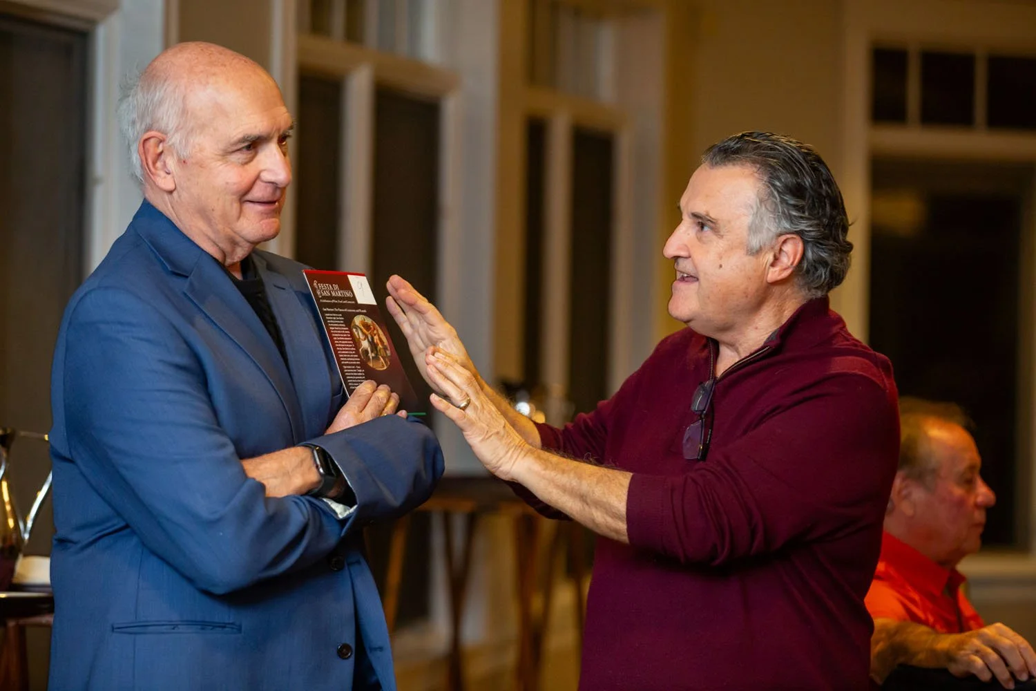 Two men are having a conversation at an indoor event. The man on the left is wearing a blue suit and is holding a brochure, and the man on the right, wearing a maroon shirt, is gesturing with his hands towards the man in the blue suit.