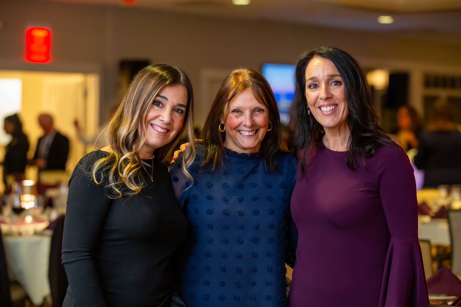 Three women smiling at a social gathering, standing closely together indoors with other people and tables in the background.