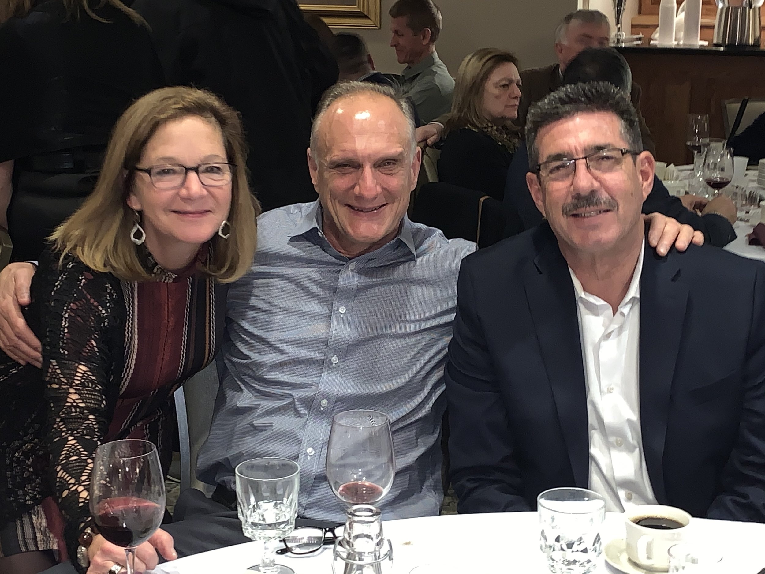 Three people sitting at a table during an event, with glasses of wine and coffee. They are smiling and appear to be enjoying themselves. The setting looks like a formal indoor gathering.
