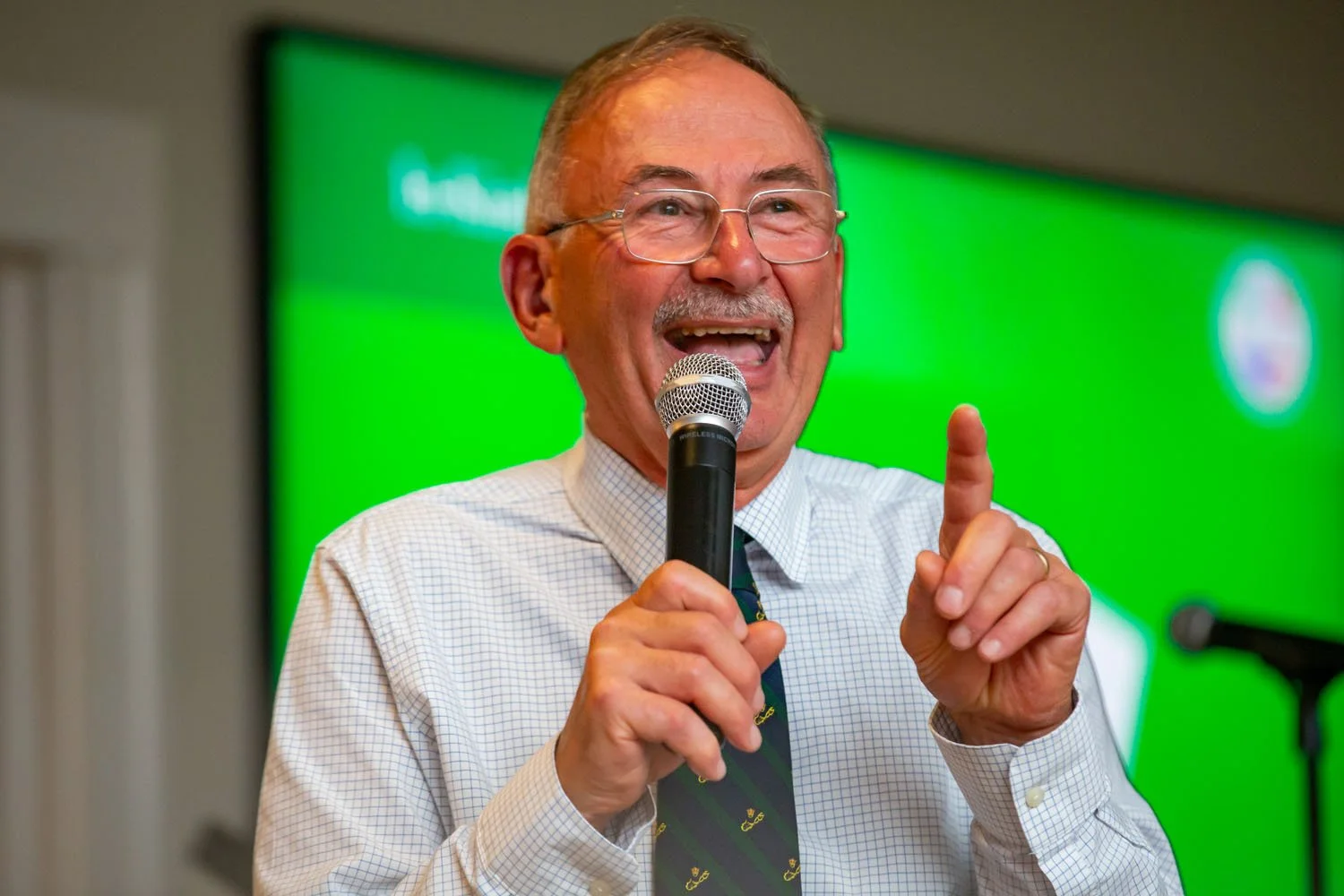 An older man with gray hair, glasses, and a mustache is speaking into a microphone during a presentation. He is wearing a white dress shirt and a green tie with a pattern. He is smiling and holding up his right index finger. The background features a