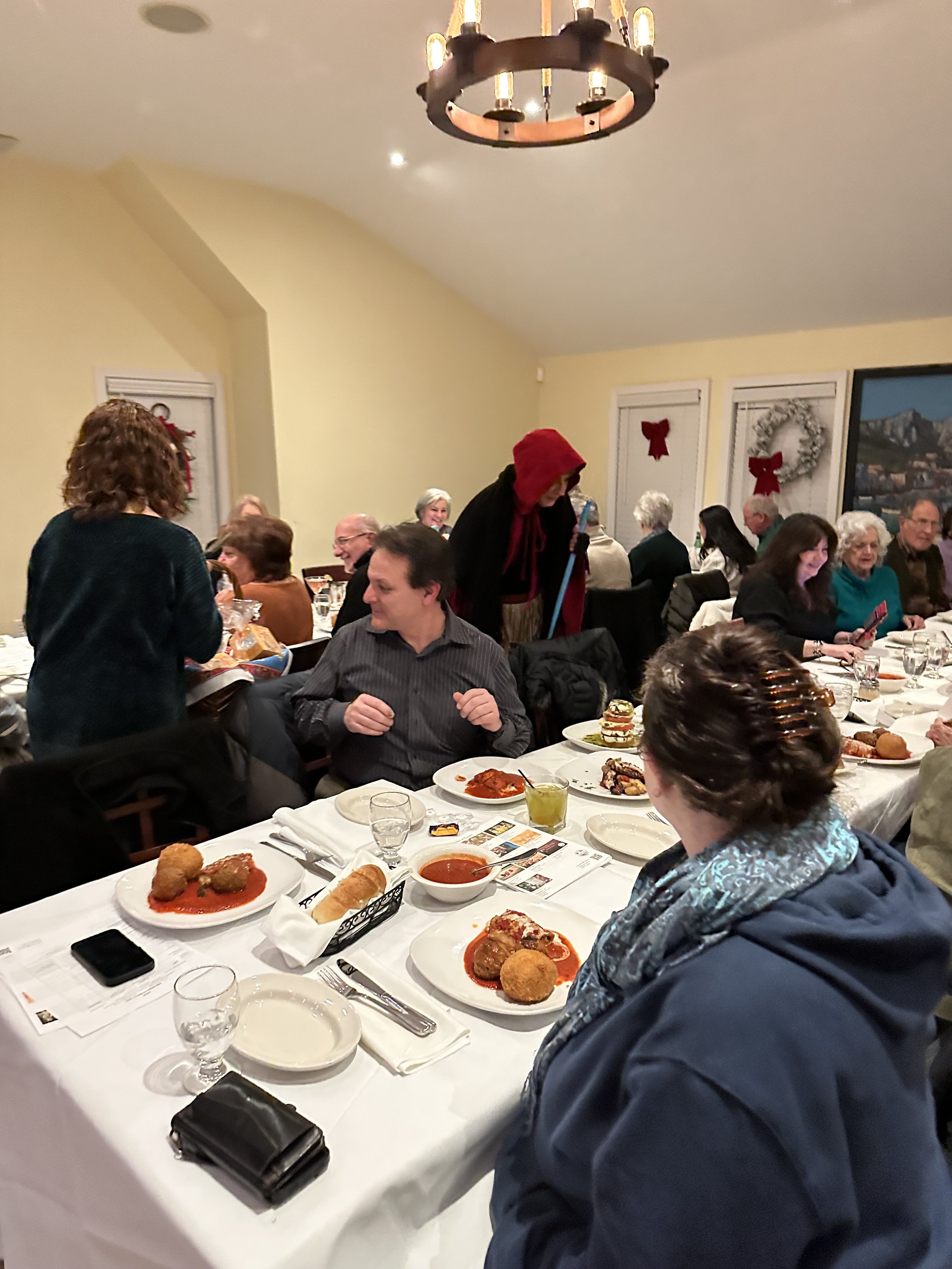 People dining at a restaurant with tables set with plates of food, including appetizers and bread, in a festive atmosphere with decorations such as wreaths and red bows on the walls.