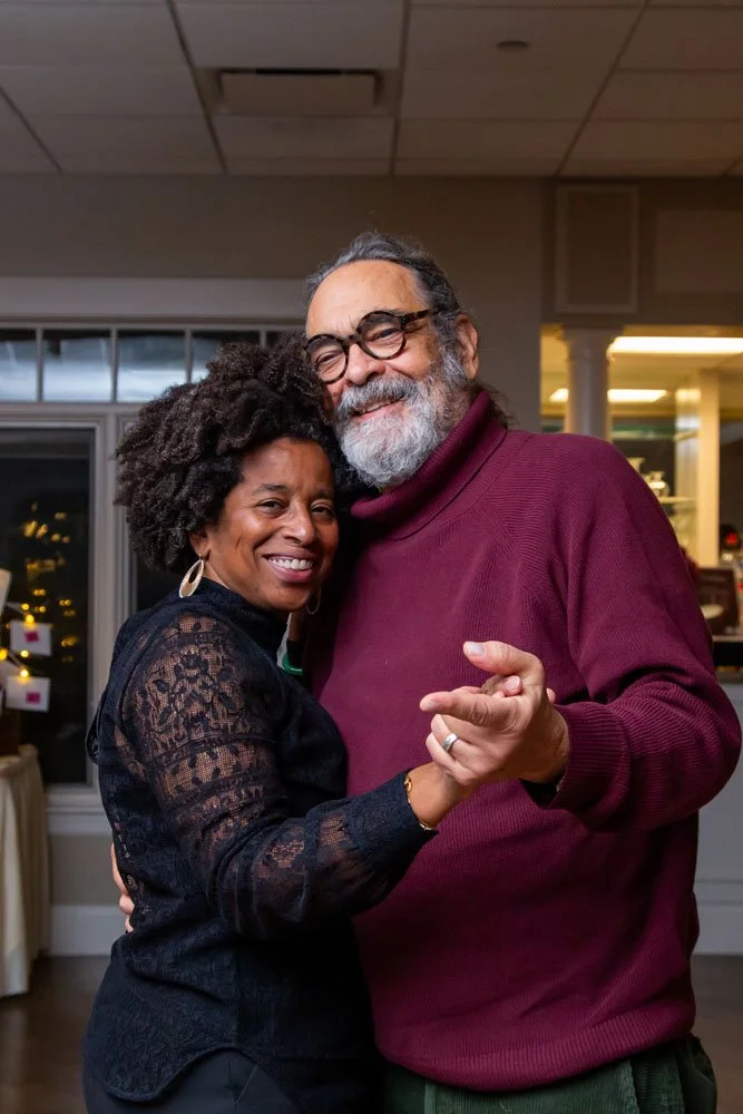 A smiling African American woman and an older man with glasses and gray beard dancing together indoors.