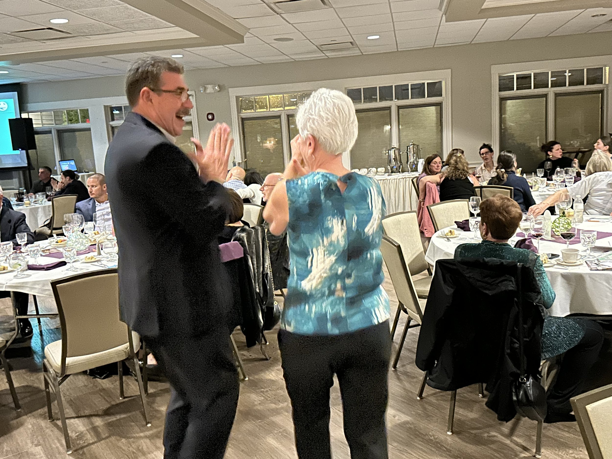Two people dancing at an indoor event, surrounded by seated guests at tables.