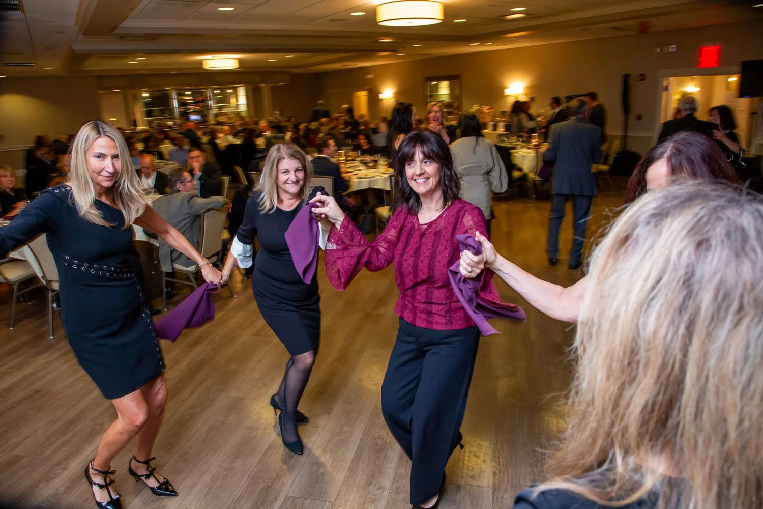 Women dancing together at a formal event in a banquet hall with tables and guests in the background.