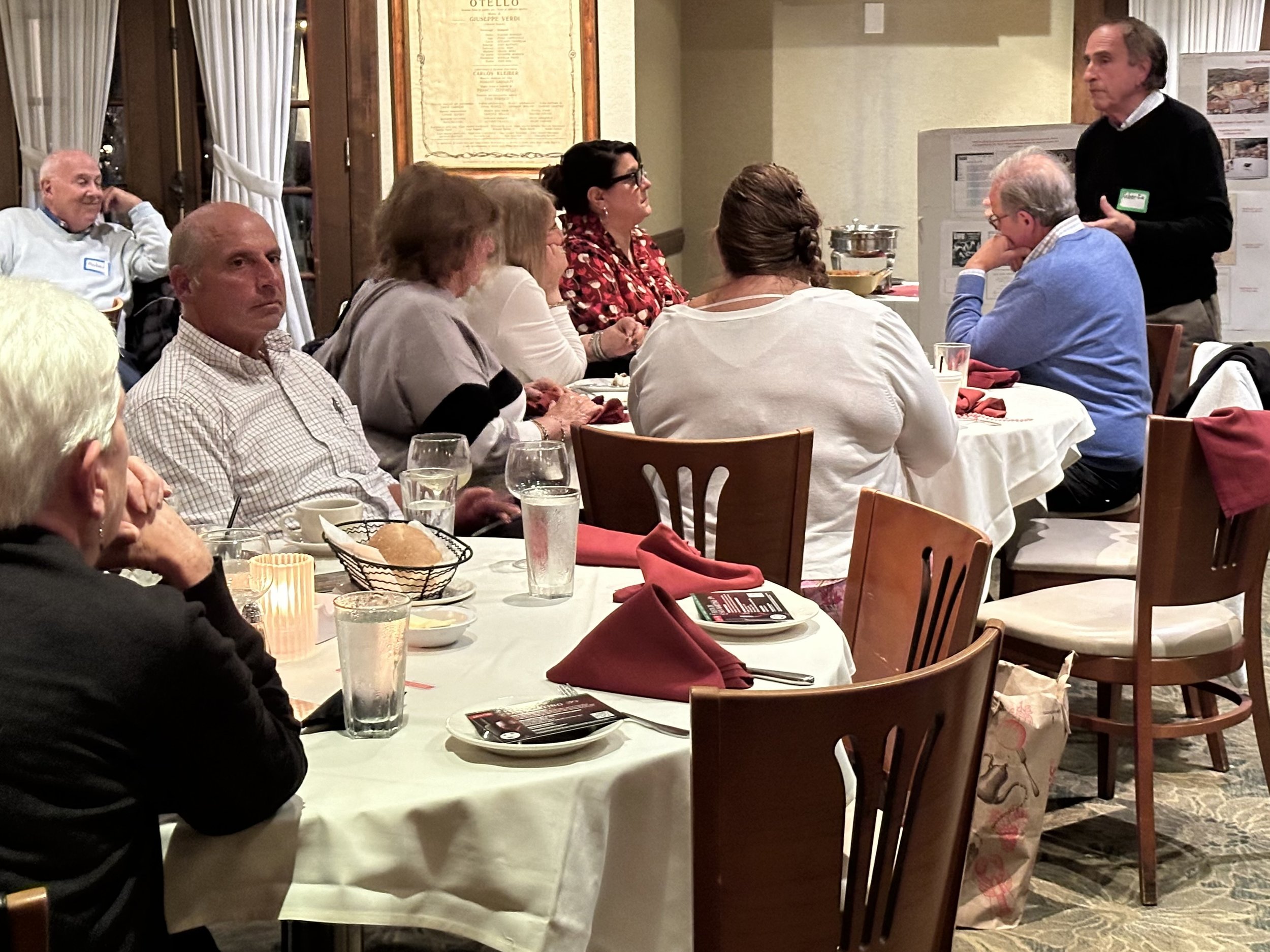 A group of people sitting around tables at a restaurant or event space, engaged in a presentation or discussion. The tables have white tablecloths and are set with glasses, plates, and napkins. One person is standing and speaking to the group.