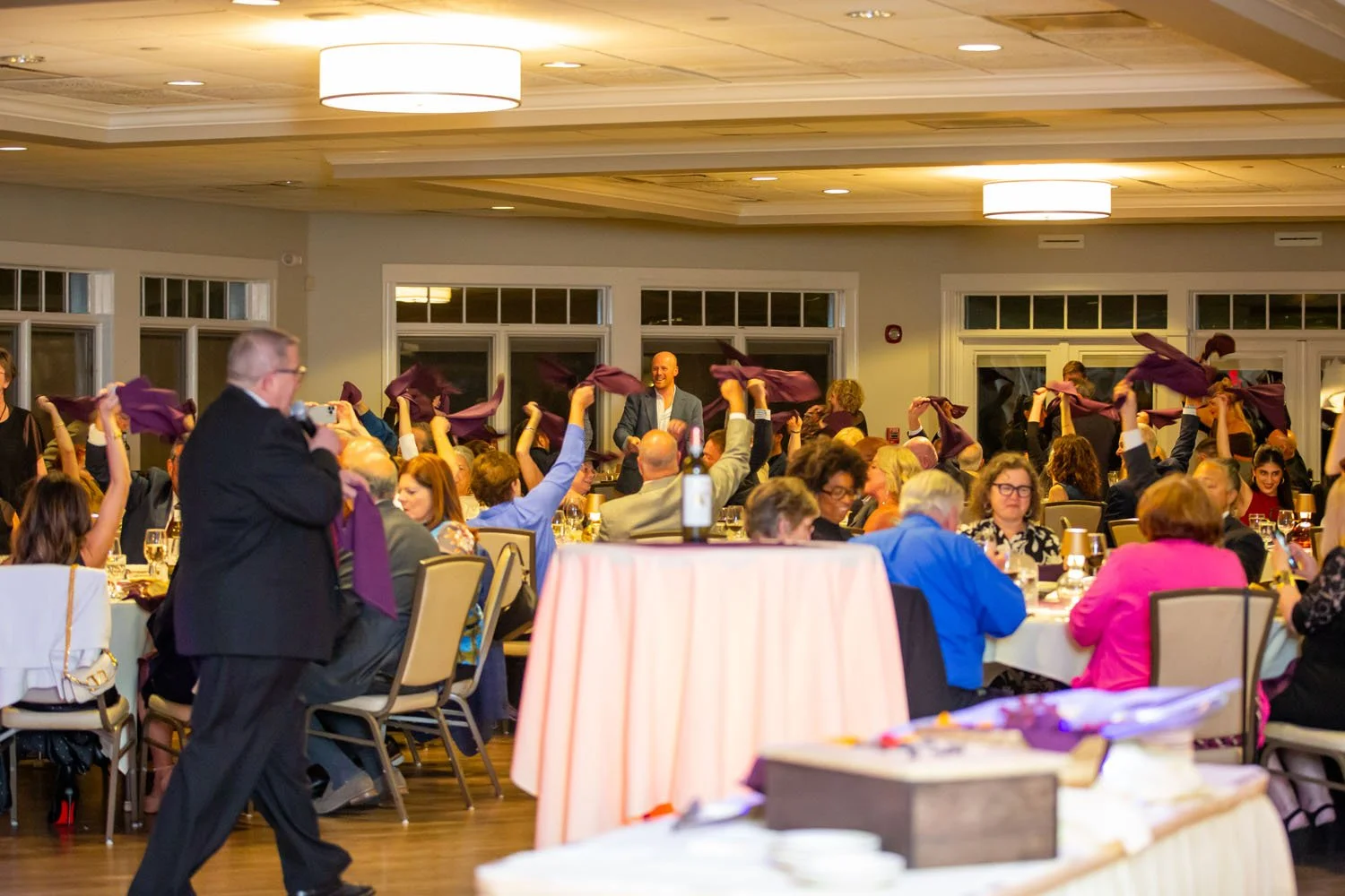 People attending an indoor event, with some raising purple napkins, and a man standing at the front speaking or presenting, in a well-lit banquet hall.