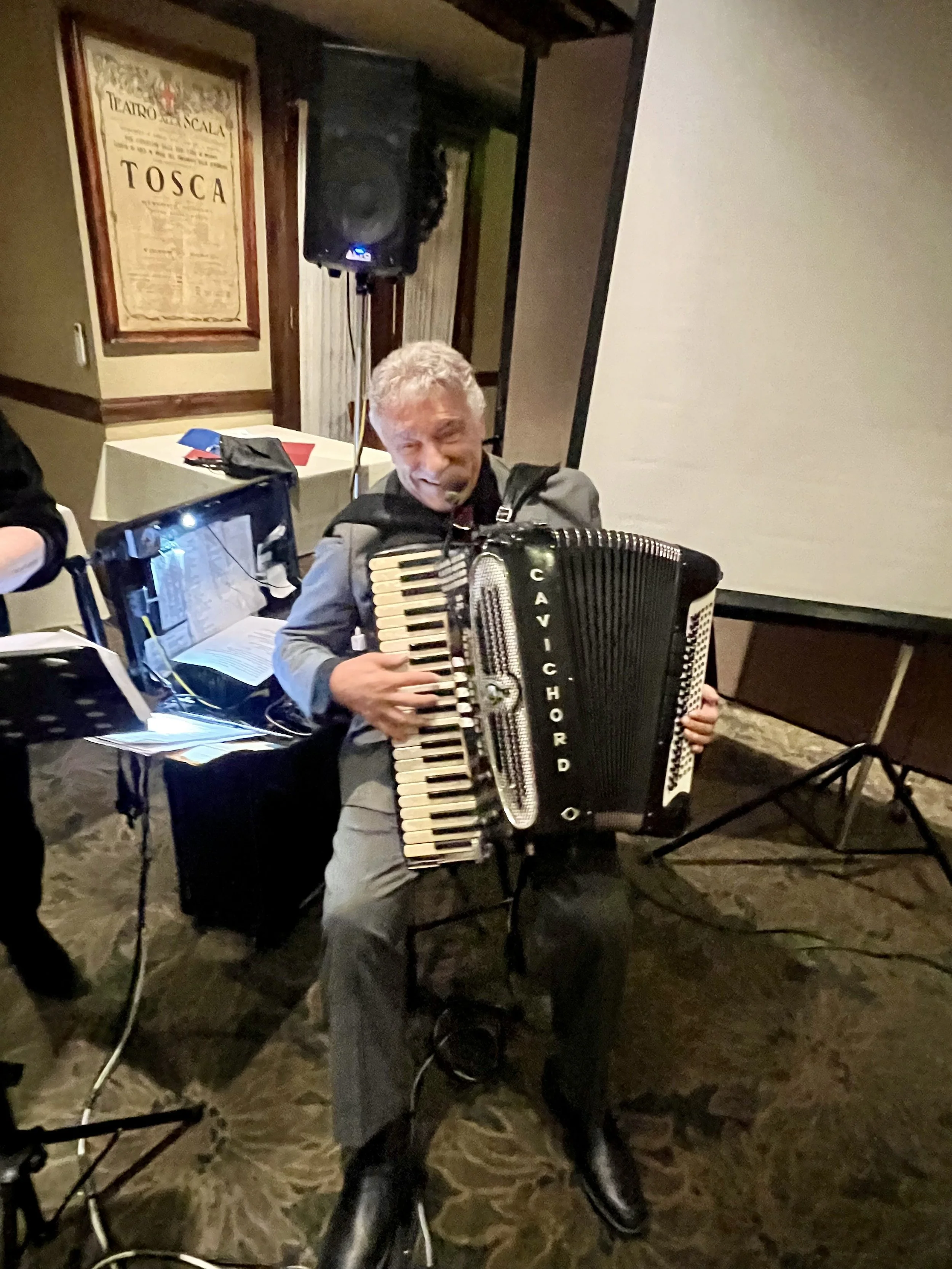 A man playing an accordion at an indoor event, with musical equipment and a projection screen nearby.