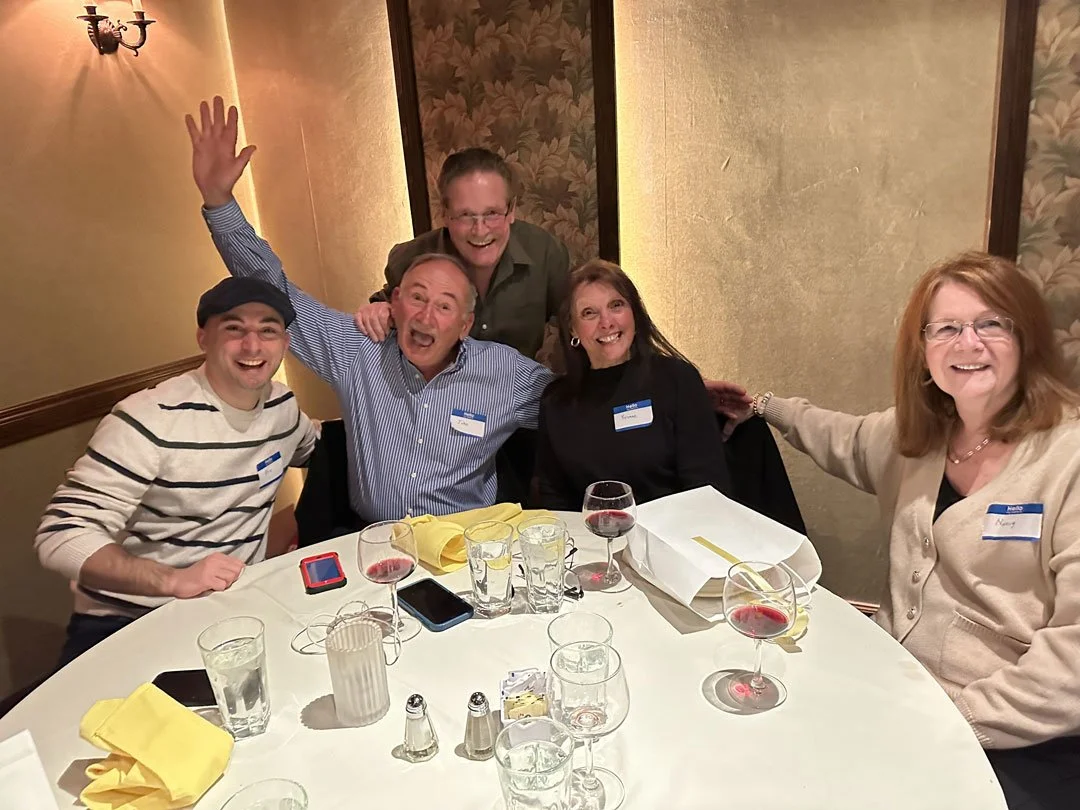 Five smiling people at a dinner table, some with glasses of red wine, in a warmly lit dining room.