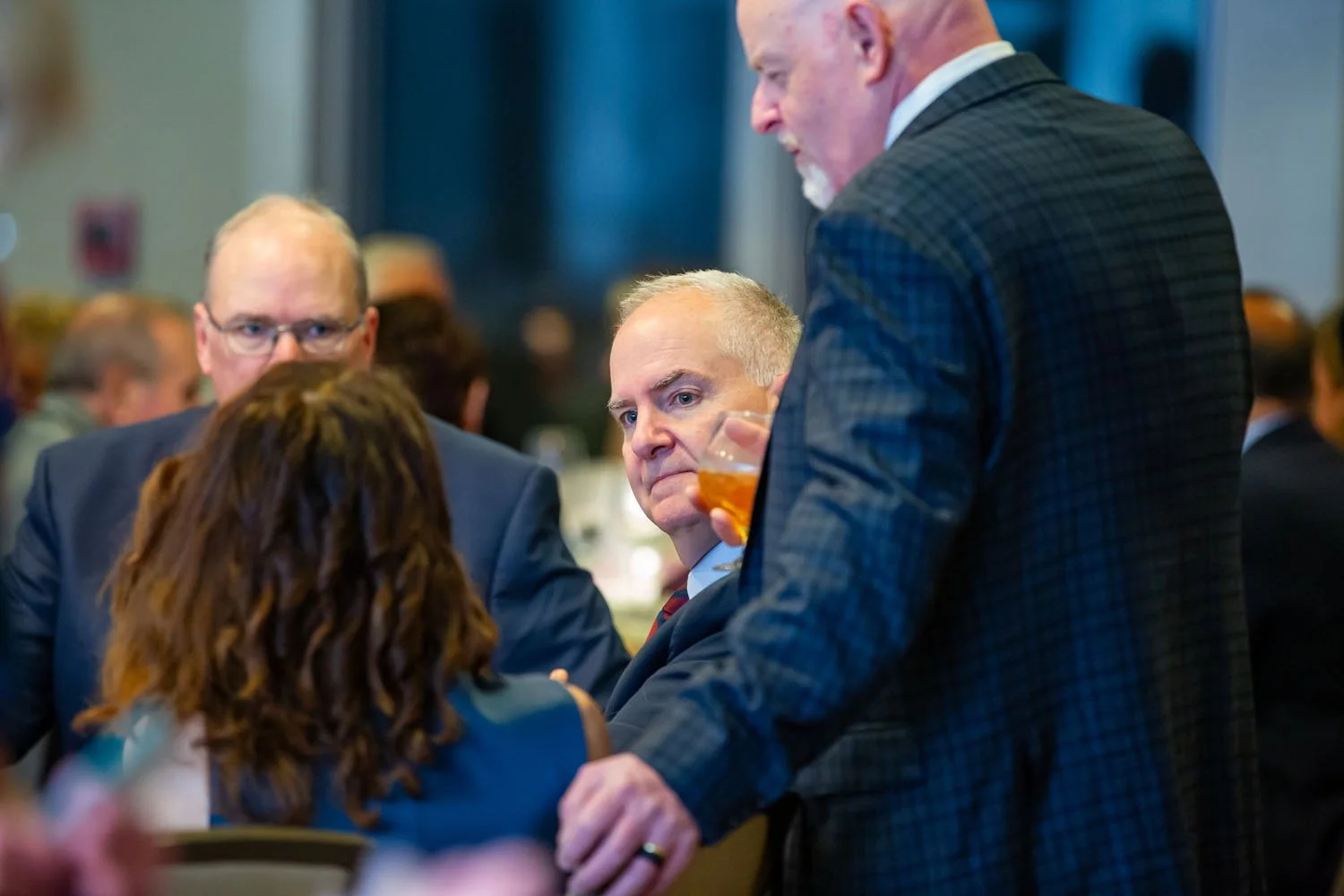 Men in suits at a social event, one holding a drink, engaging in conversation.