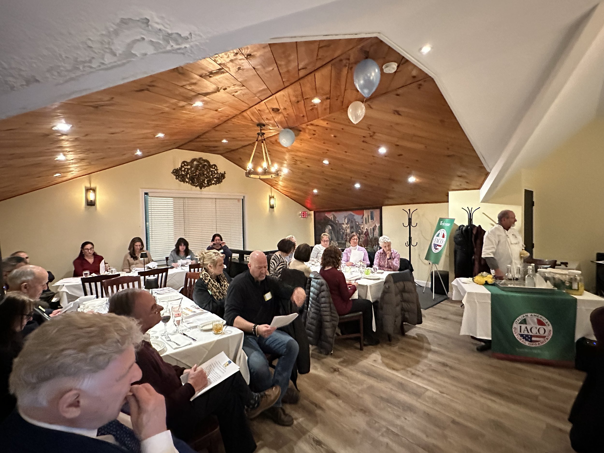 A group of people seated at tables in a warmly lit restaurant or dining room, attending a presentation or meeting. A speaker is standing near a table with a sign labeled 'IACO'. The room has a wooden ceiling, and decorations include balloons and wall