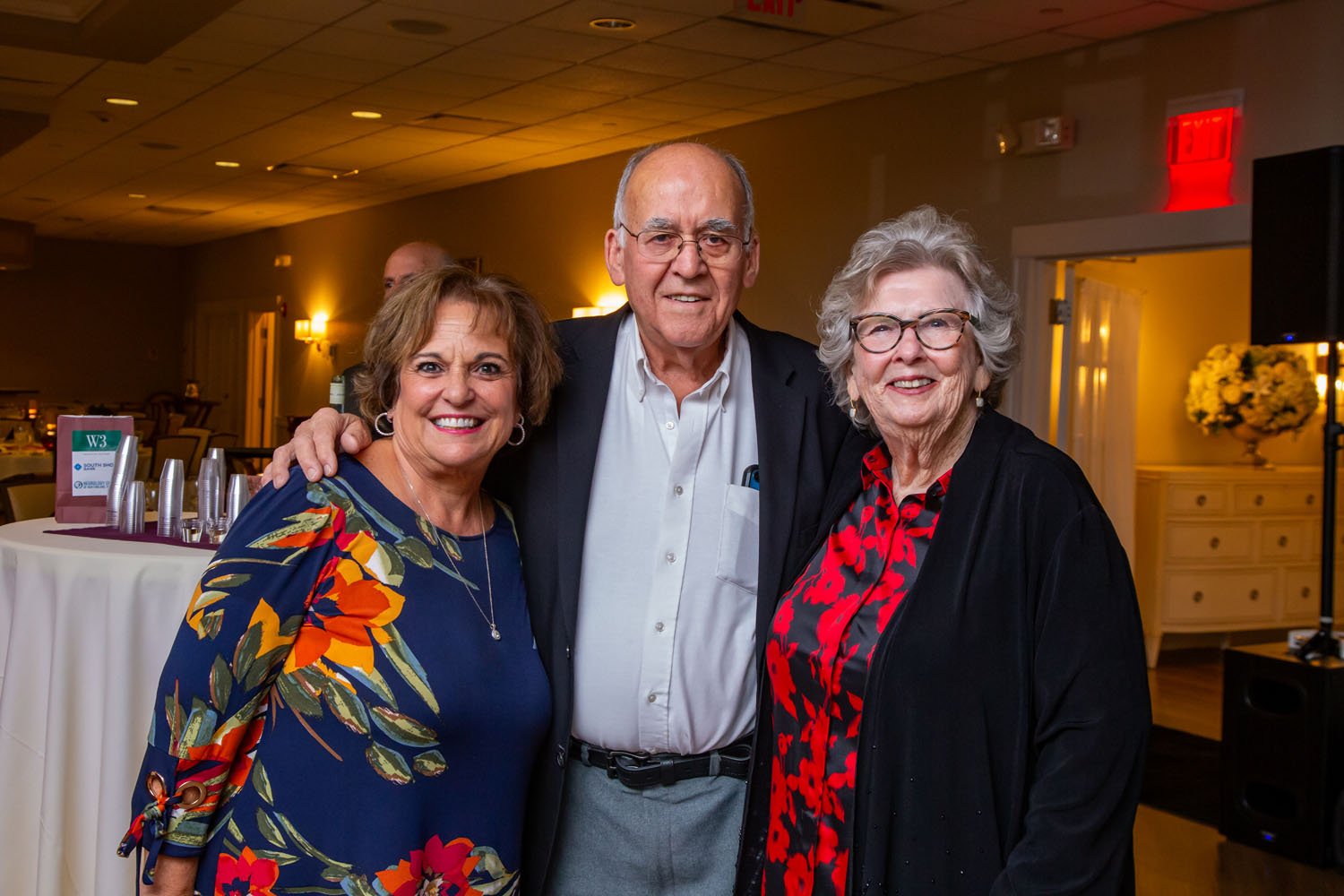 Three smiling seniors, two women and one man, at a social event in a banquet hall.