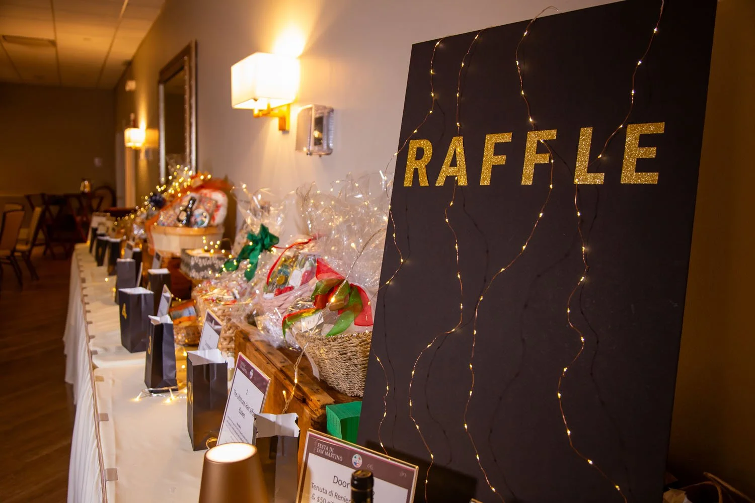 A table displaying raffle prizes with a black sign that says 'RAFFLE' in gold glitter letters, decorated with string lights, in an indoor event setting.