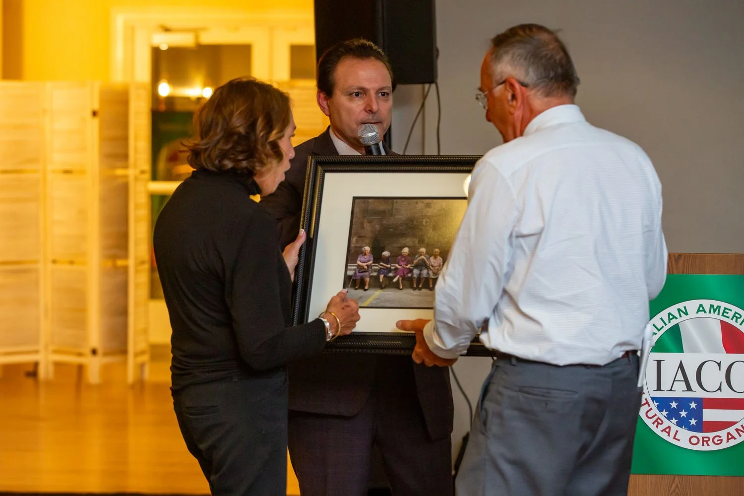 Three individuals at an event, two are presenting a framed photo to a man. The woman on the left holds the frame, the man on the right receives it, and another man in the middle speaks into a microphone. The framed photo shows a group of elderly wome