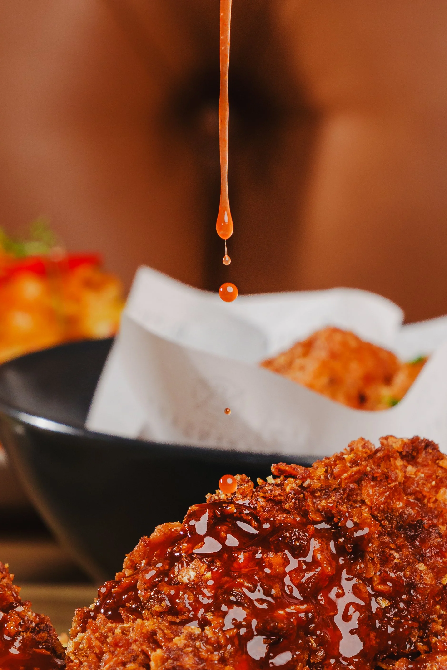 Close-up of a savory breaded fried meat patty topped with barbecue sauce, with droplets of sauce falling from above, set against a blurred background with some food items.