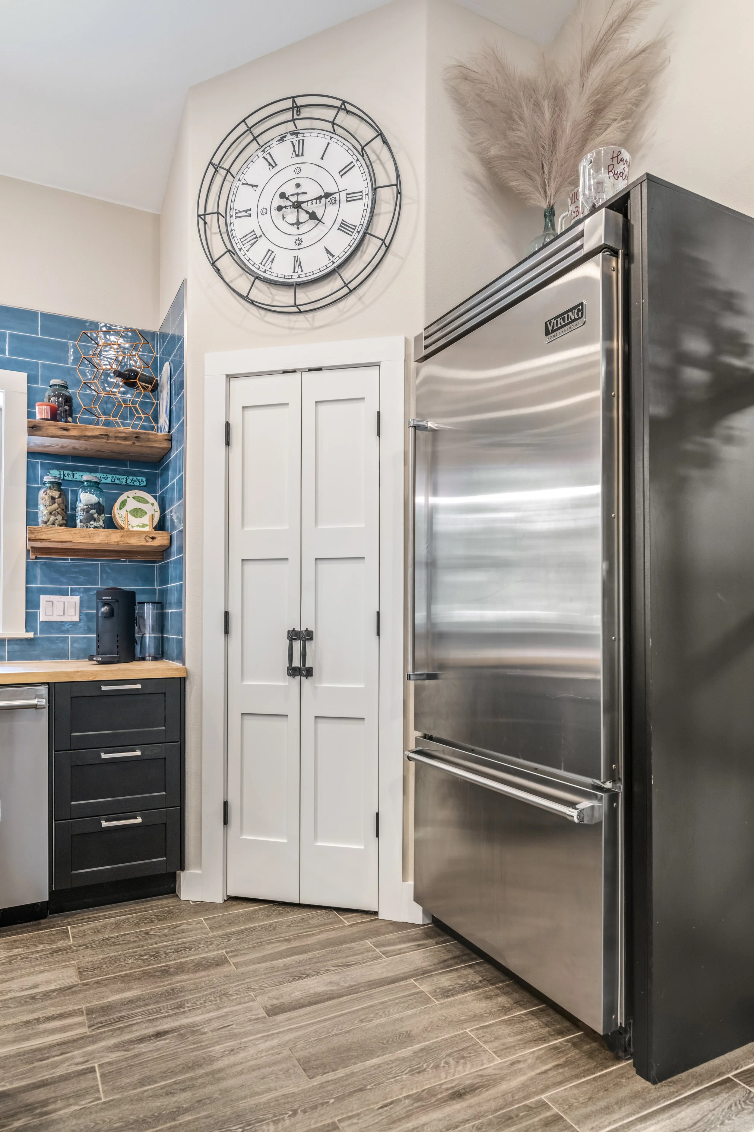 Modern kitchen corner with blue tiled wall, wooden shelves holding jars, and a stainless steel refrigerator. A large decorative clock hangs on the wall above a white pantry door.