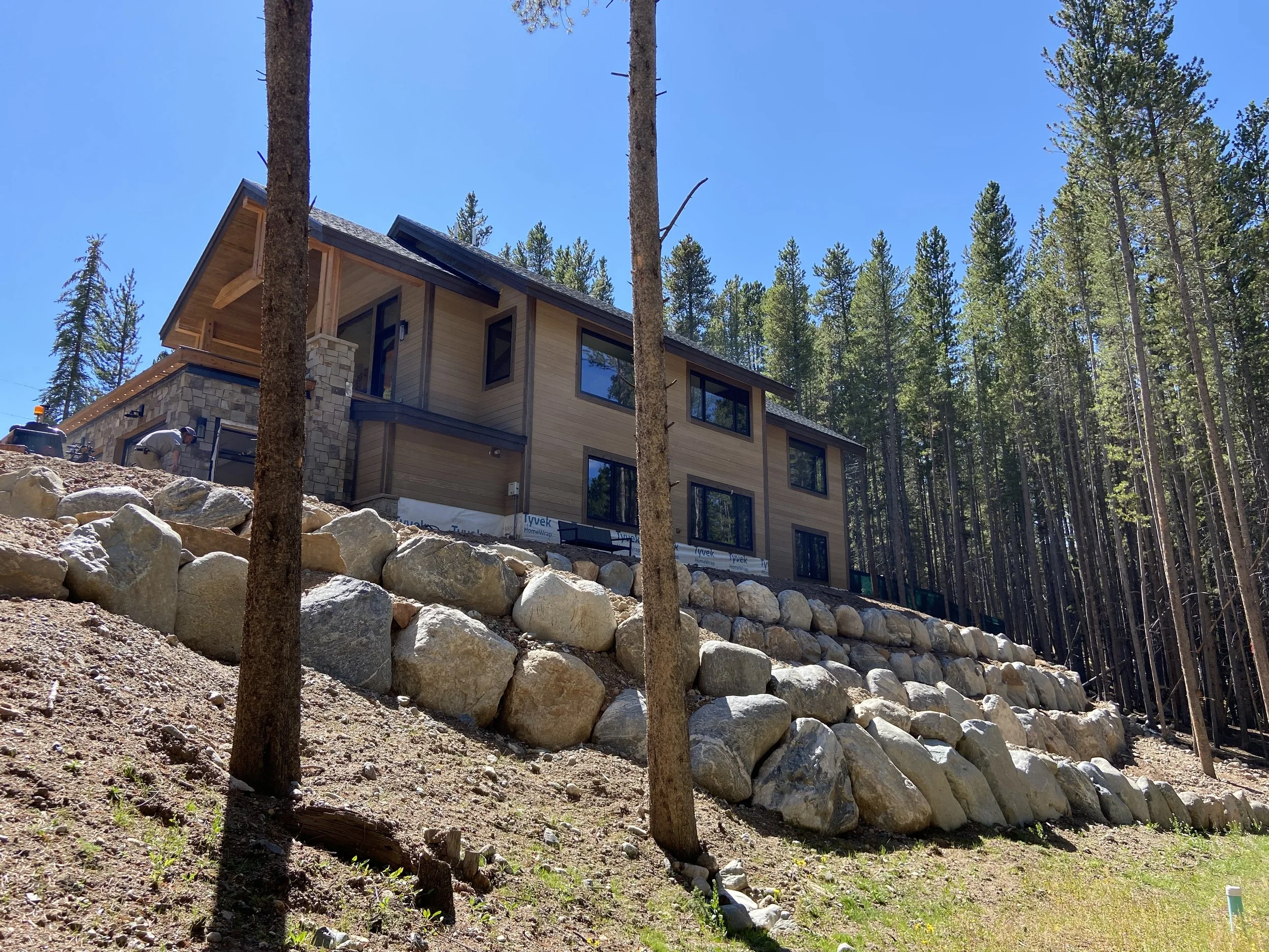 Two-story modern house with large windows and wooden siding, surrounded by a forest of tall pine trees. The house is built on a slope with a stone retaining wall. Clear blue sky in the background.