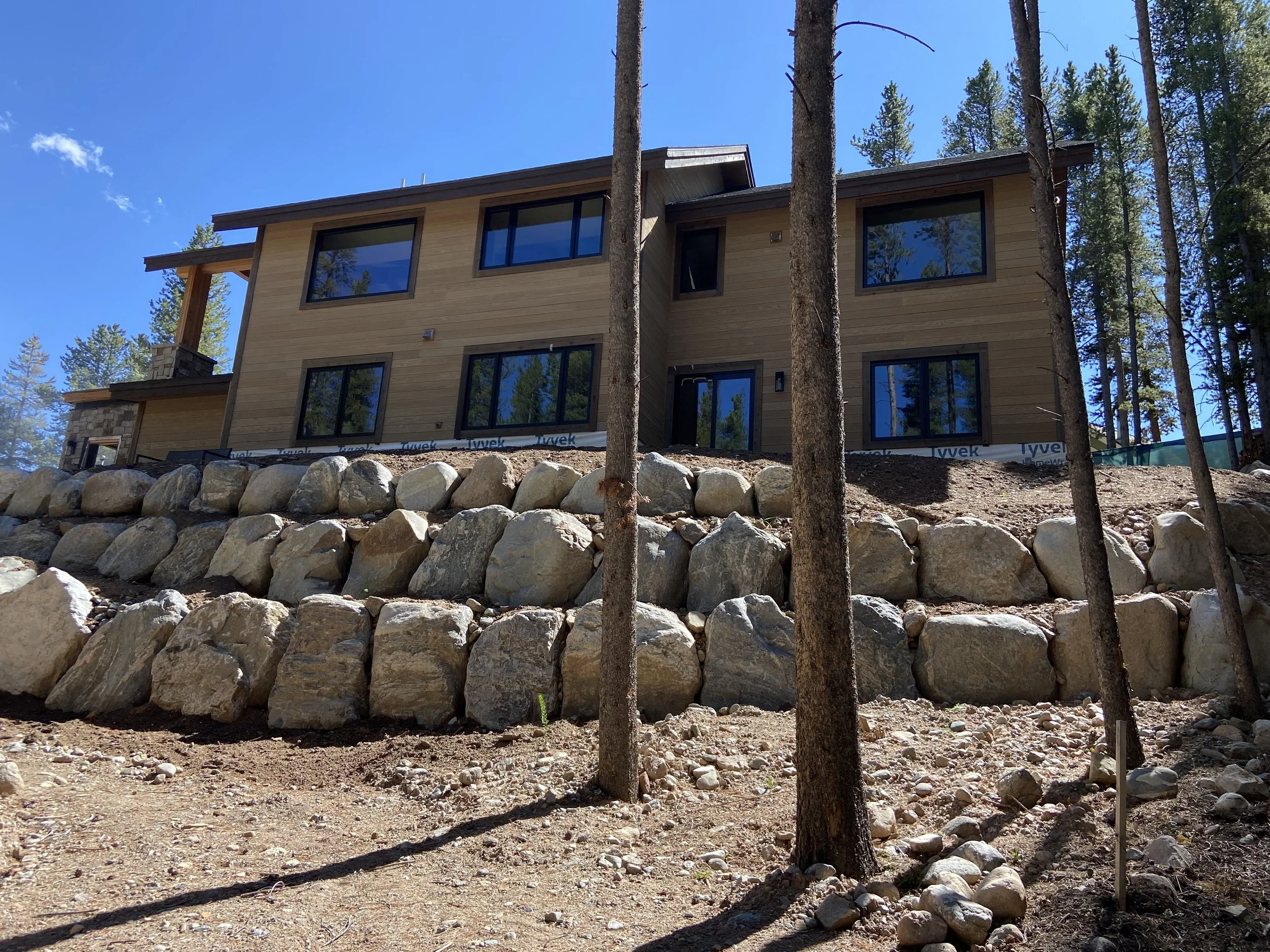 Modern house under construction on a rocky slope surrounded by trees.