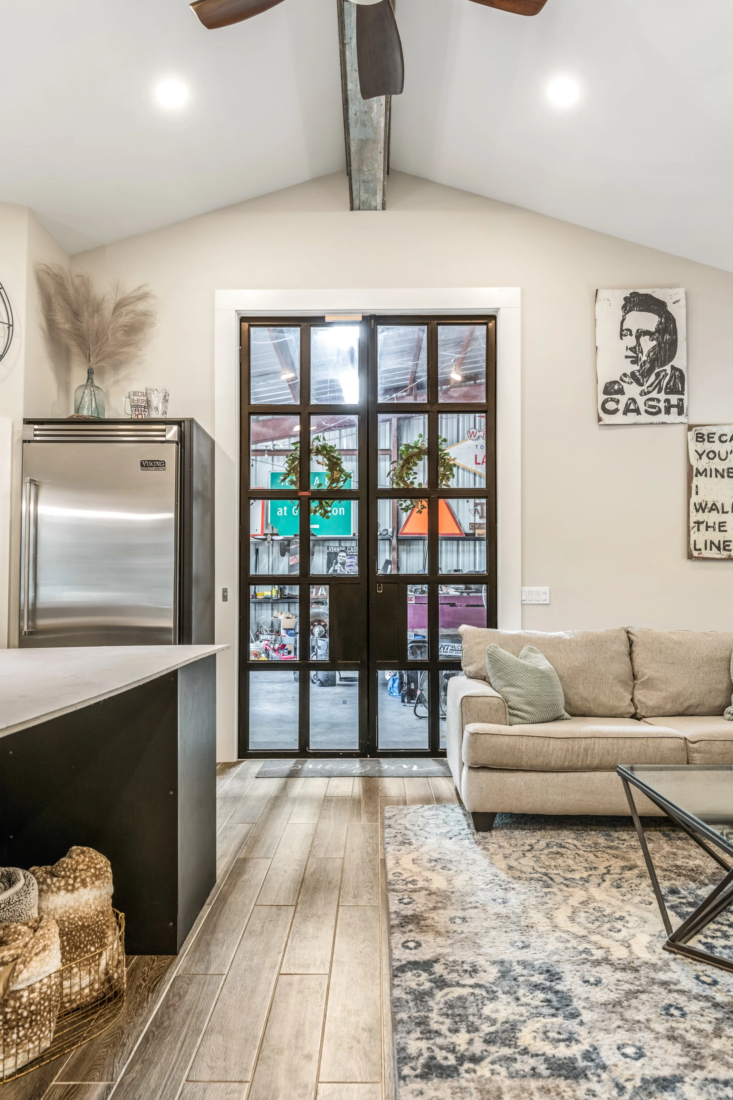 Modern living room interior with glass double doors, beige sofa, glass coffee table, patterned rug, wall art, stainless steel refrigerator, and exposed ceiling fan.
