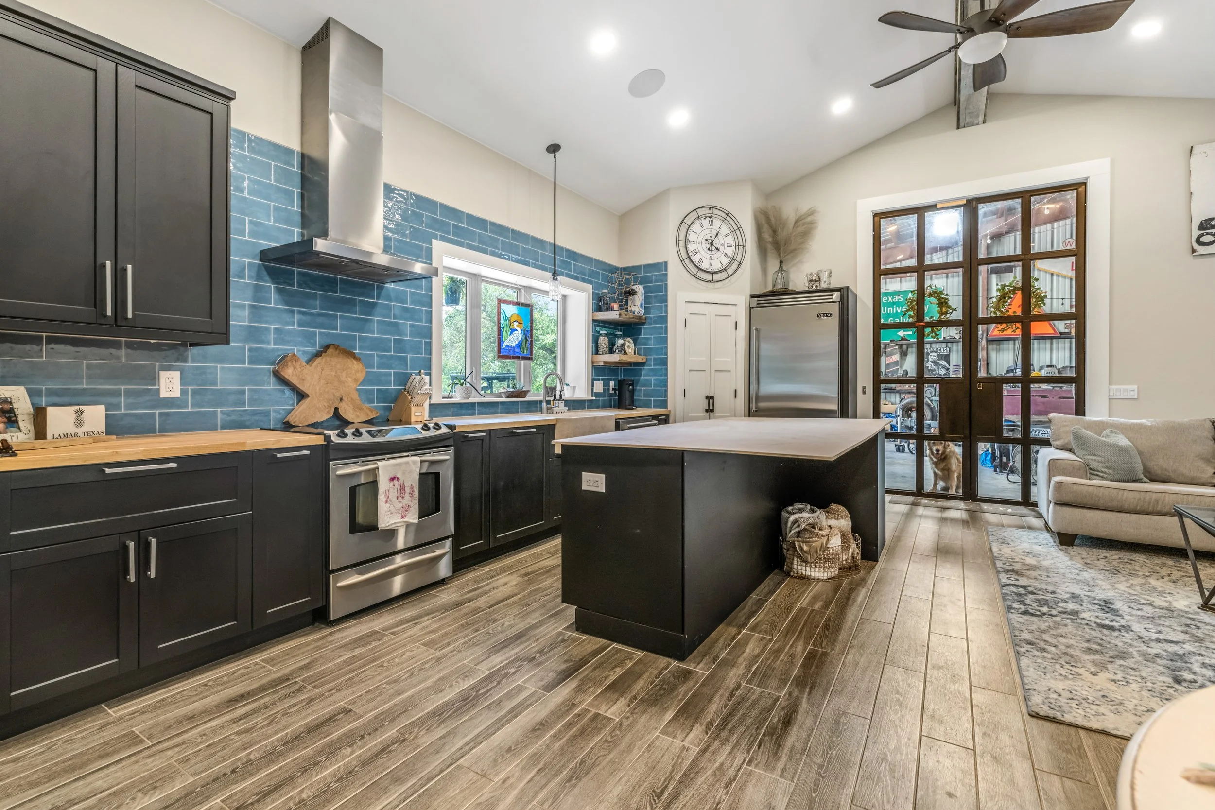 Modern kitchen with dark cabinets, wooden countertops, and a blue tile backsplash. Features include a stainless steel stove and range hood, large island with wicker baskets, and a silver fridge. There are large windows with stained glass detail, a cl