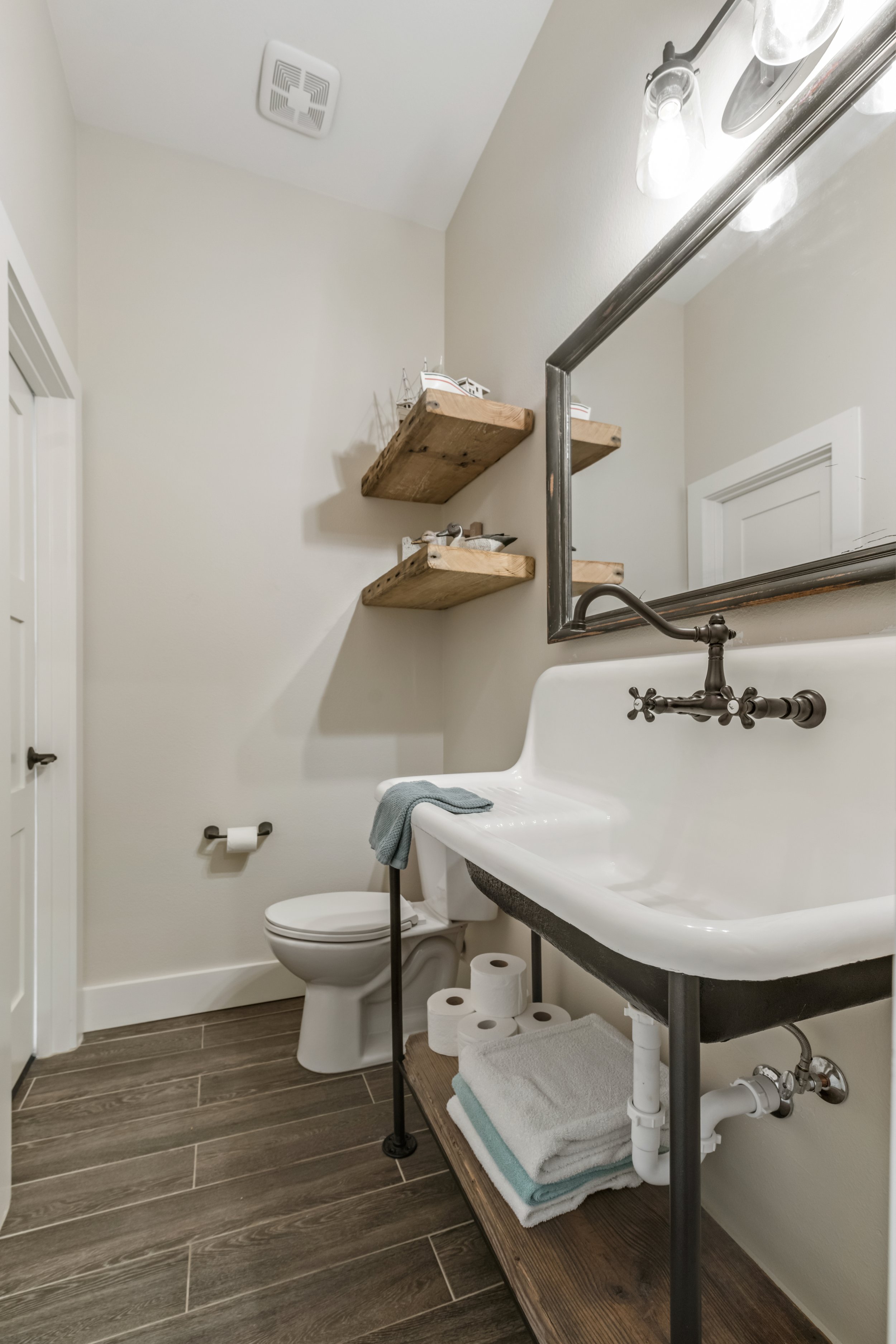 Bathroom with white wall-mounted sink, toilet, wooden shelves, towels, and toilet paper.