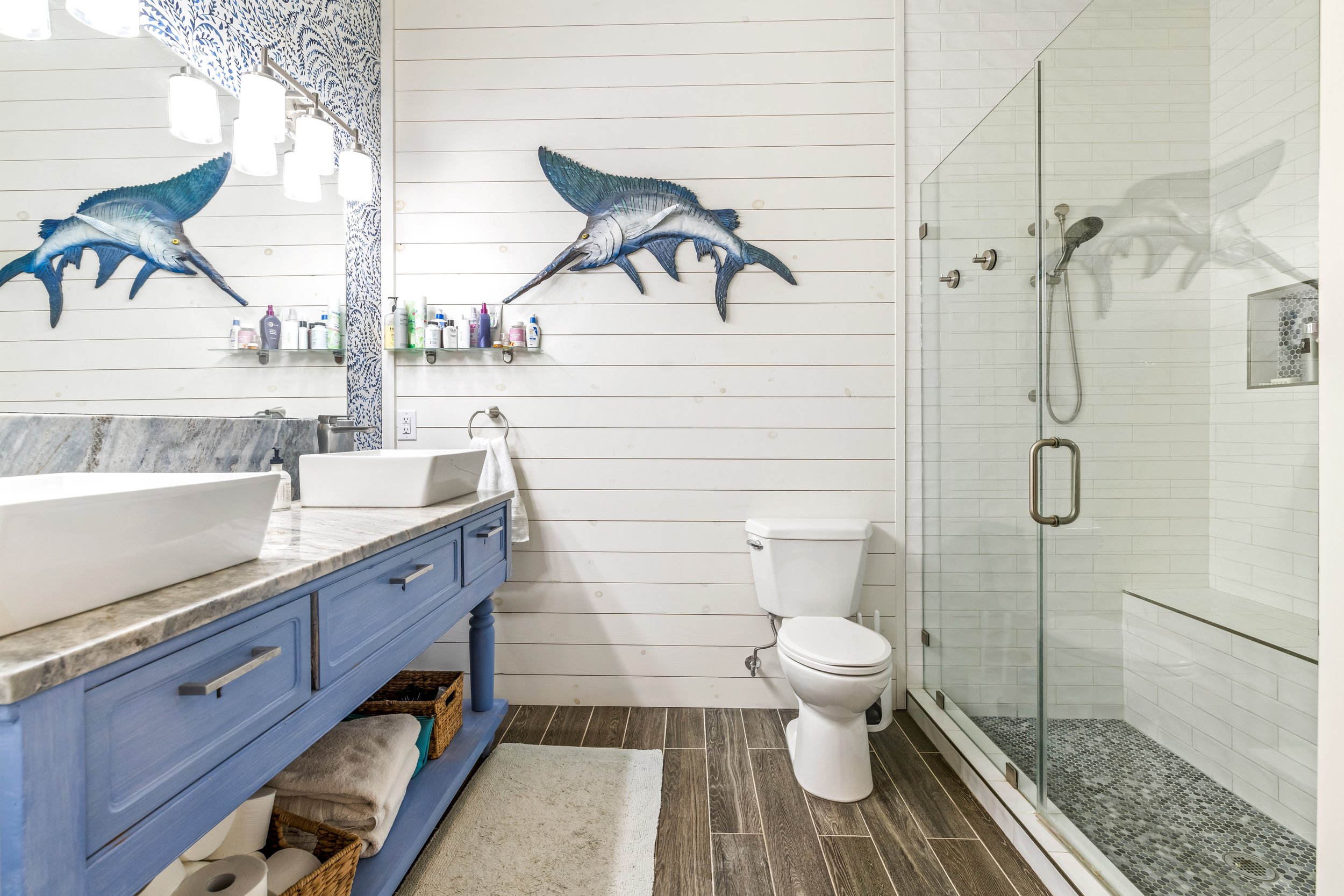 Beach-themed bathroom with blue vanity, dual sinks, mirror, shower, and a marlin wall decoration.