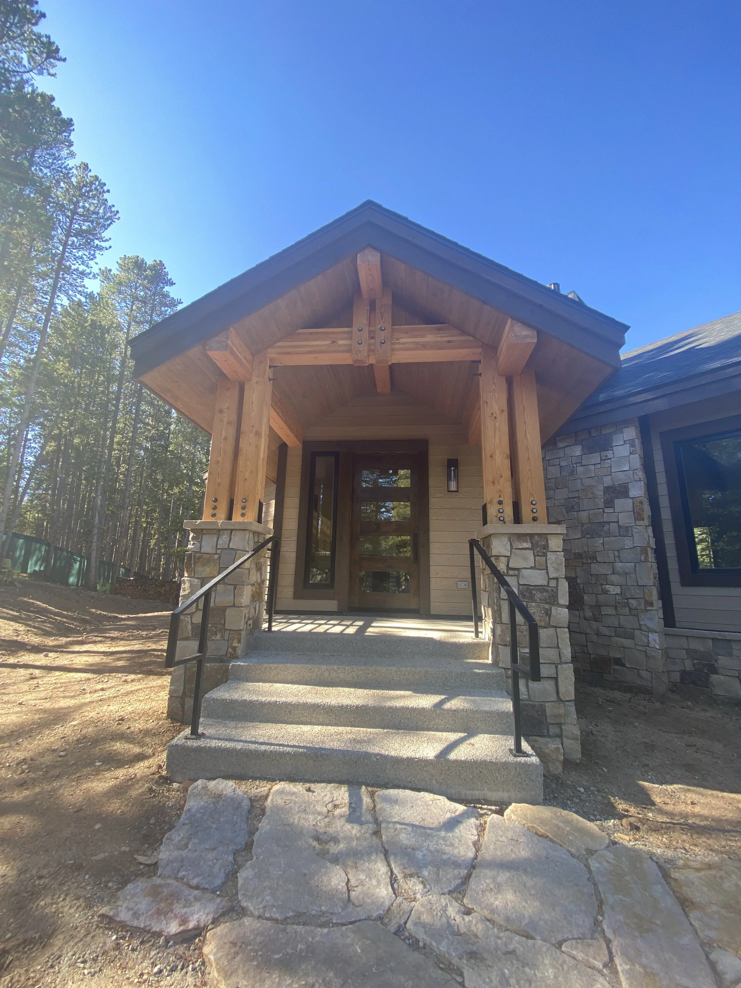 Front entrance of a house with a wooden porch, stone steps, and metal handrails, surrounded by trees under a blue sky.