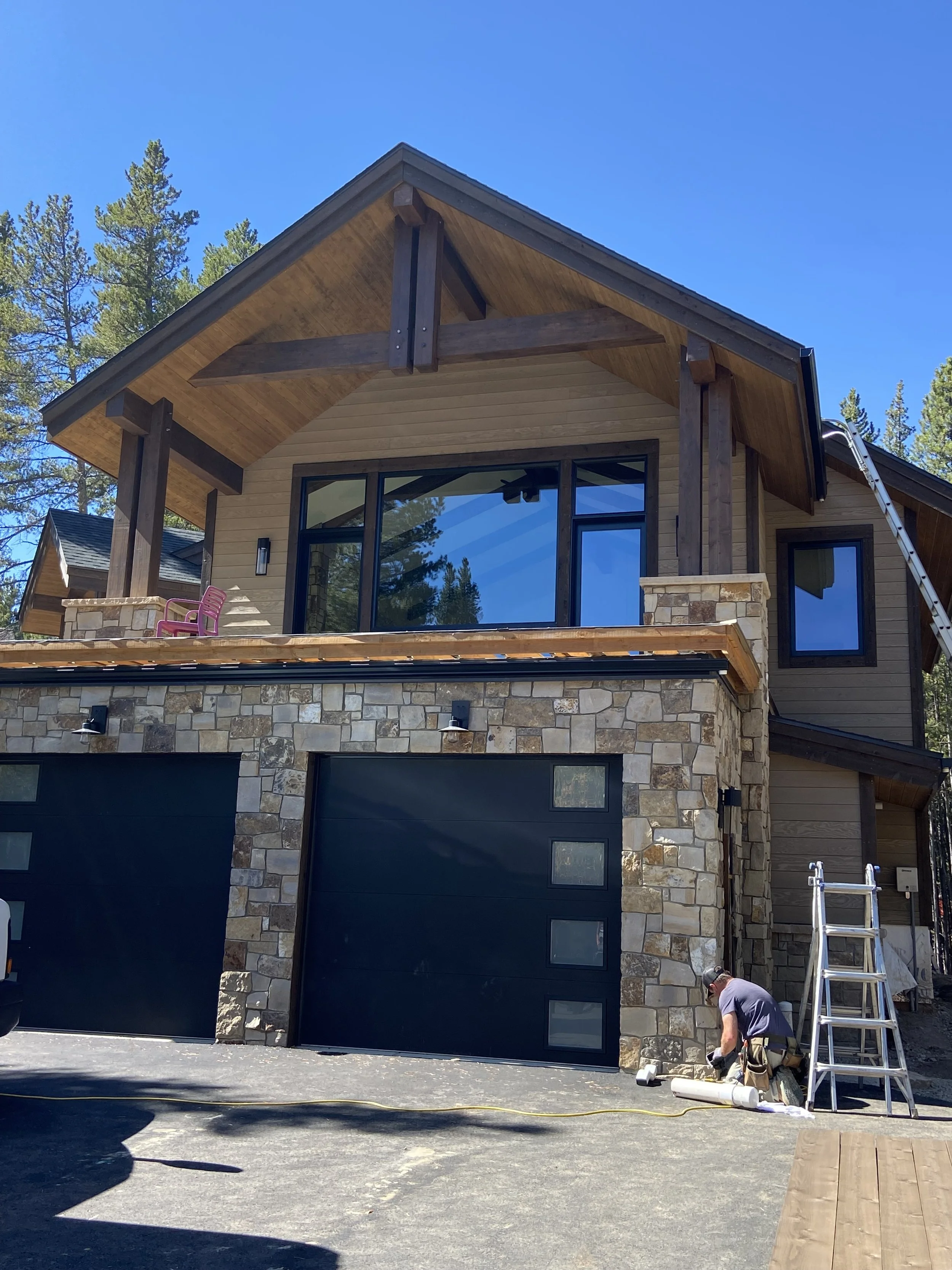 Modern two-story house with stone and wood exterior, featuring two black garage doors. A person is working near the garage, and a ladder is leaned against the wall. The house is set against a backdrop of trees under a clear blue sky.