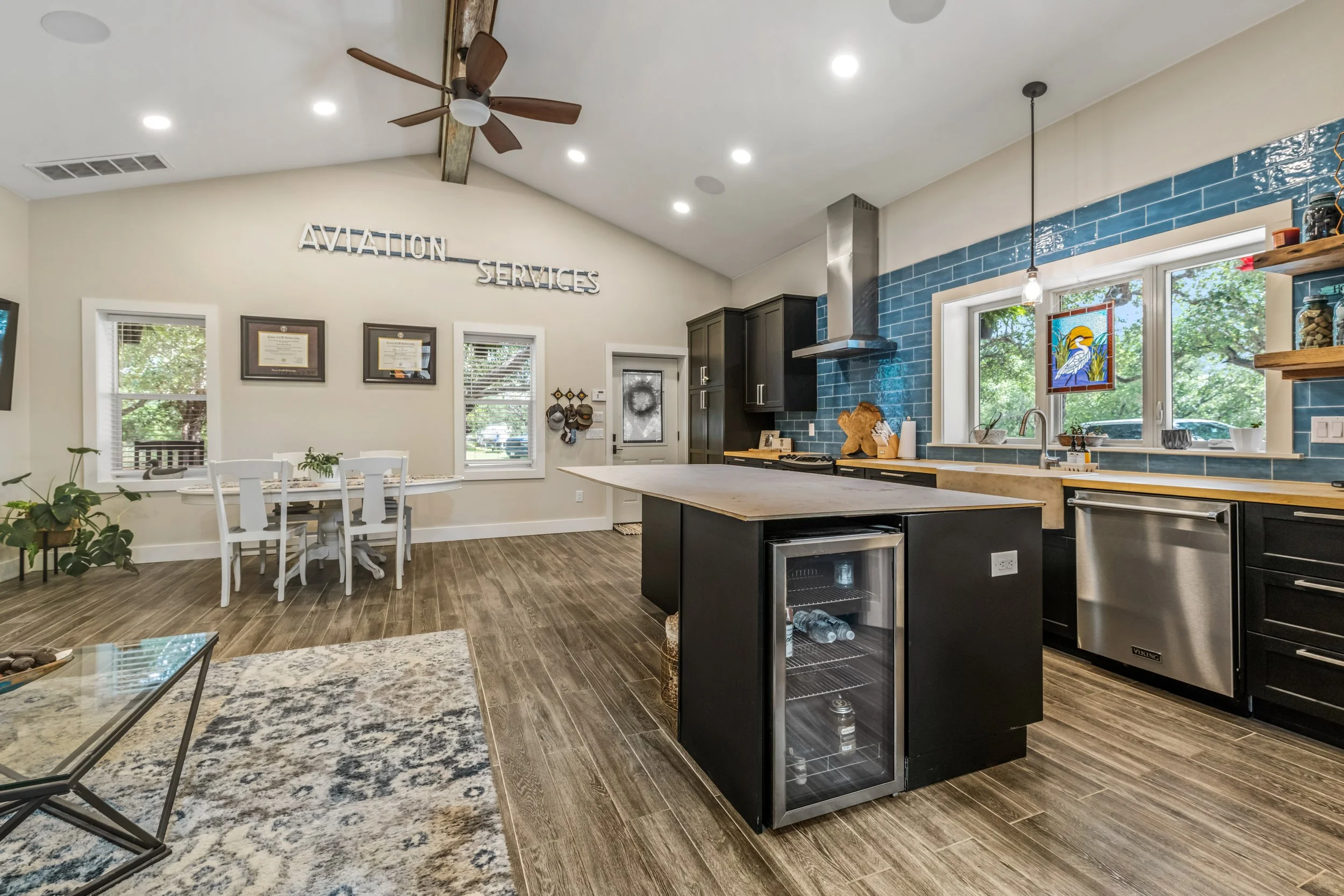 Modern kitchen and dining area with black cabinets, blue tile backsplash, center island with glass-front fridge, ceiling fan, wall art, and wooden dining table.