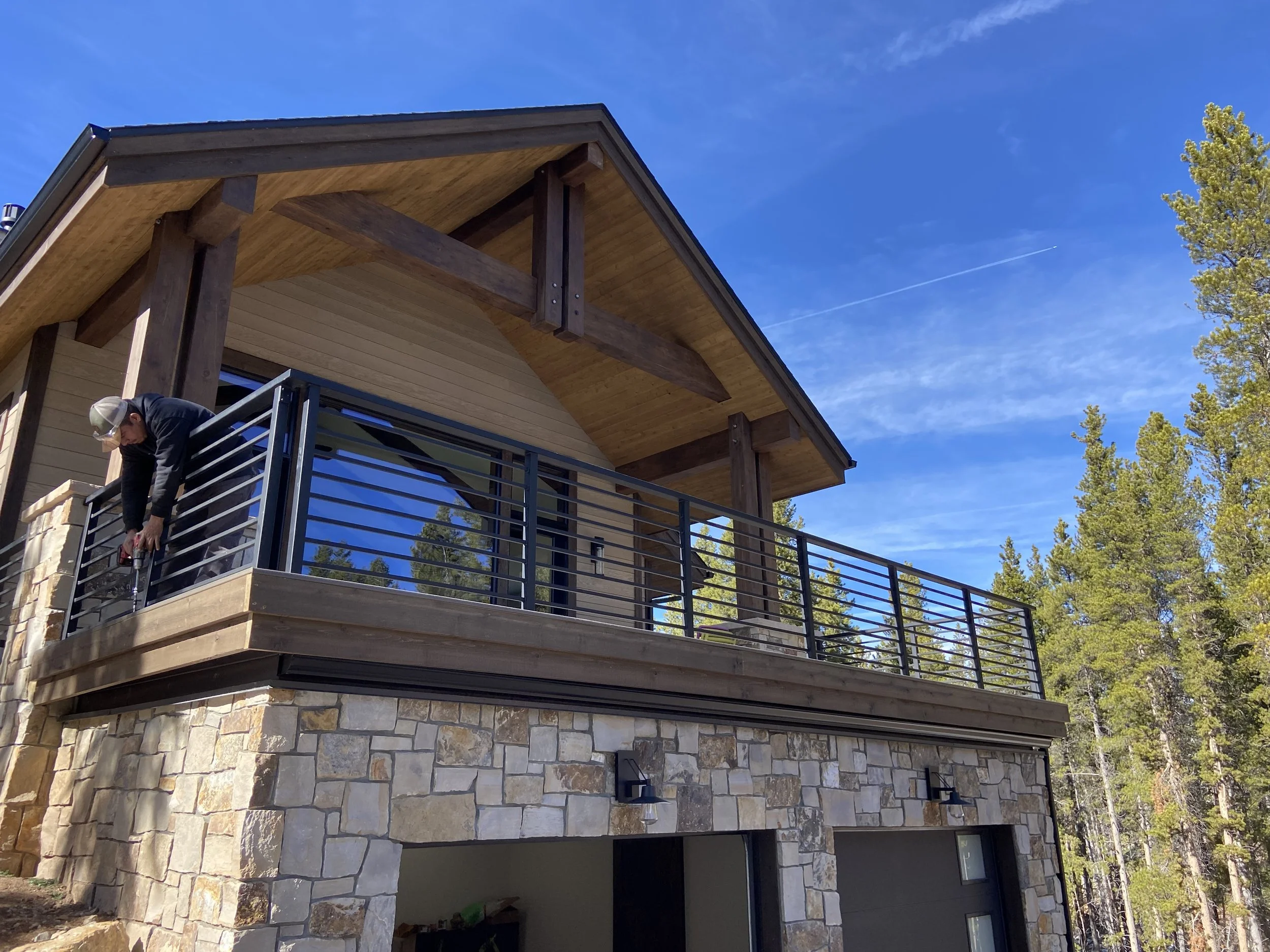 House with stone exterior and wooden balcony overlooking a forest, person working on railing.