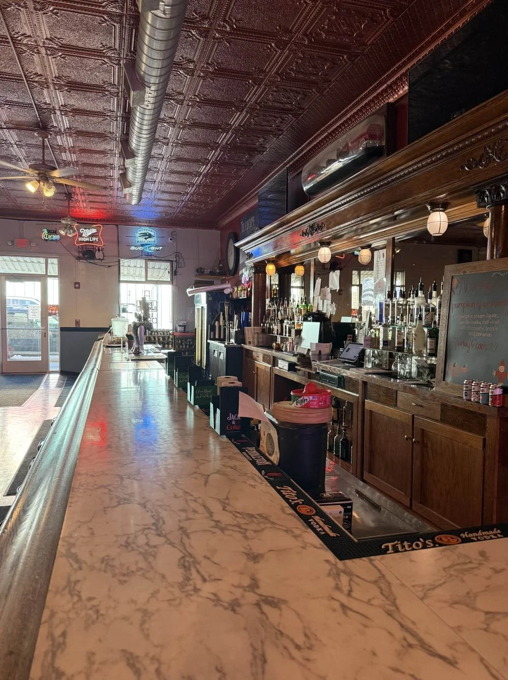 Empty bar with wooden counter and back wall featuring liquor bottles, illuminated paper lanterns, neon signs, and large windows letting in natural light.