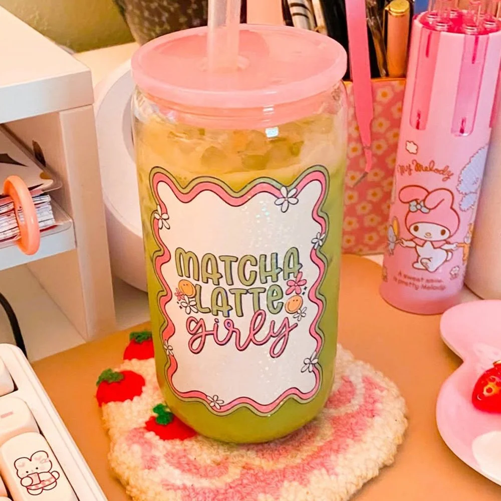 A clear plastic jar with a pink lid and straw, labeled 'Matcha Latte Girl' with flowers and strawberries, placed on a textured cloth on a desk. Nearby are pink Hello Kitty themed items, a strawberry-shaped object, and other stationery.