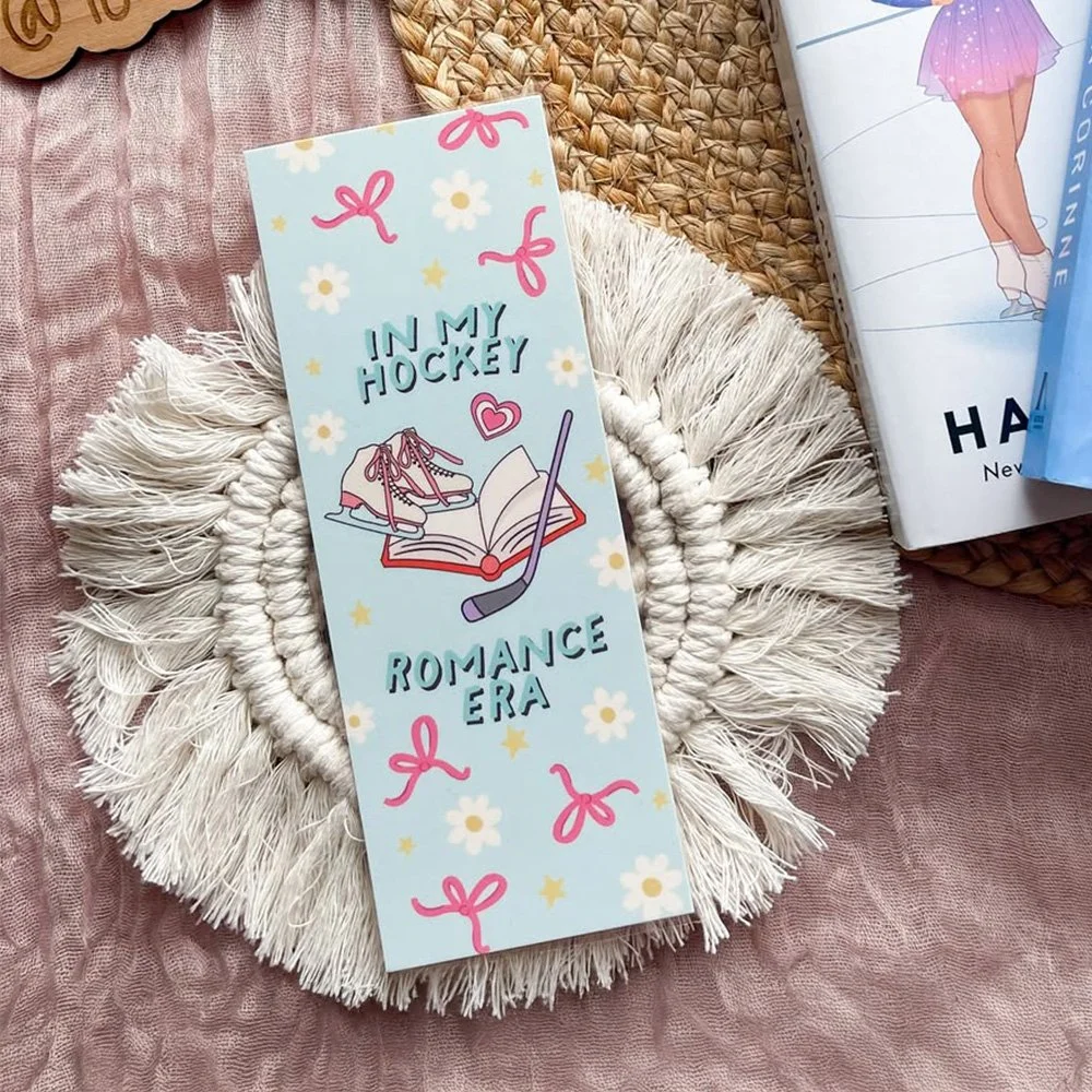 Decorative card with ice skates, hockey stick, and open book illustration, titled 'In My Hockey Romance Era,' placed on a round, fringed, white woven placemat on a pink textured surface, with other books nearby.