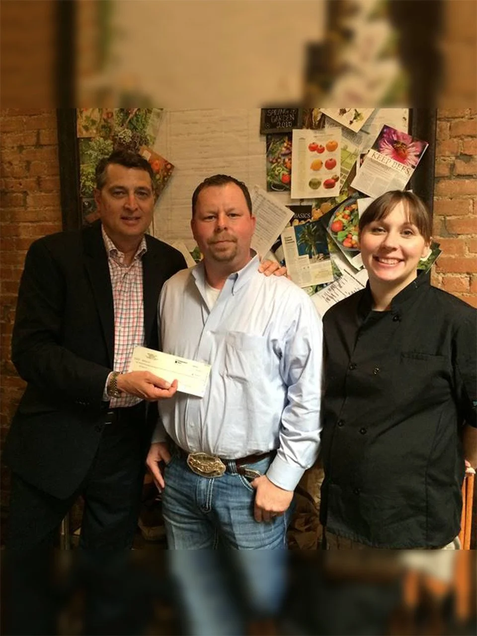 Three people standing in front of a bulletin board, one man holding a check, another man in a white shirt, and a woman in a black chef coat, all smiling.