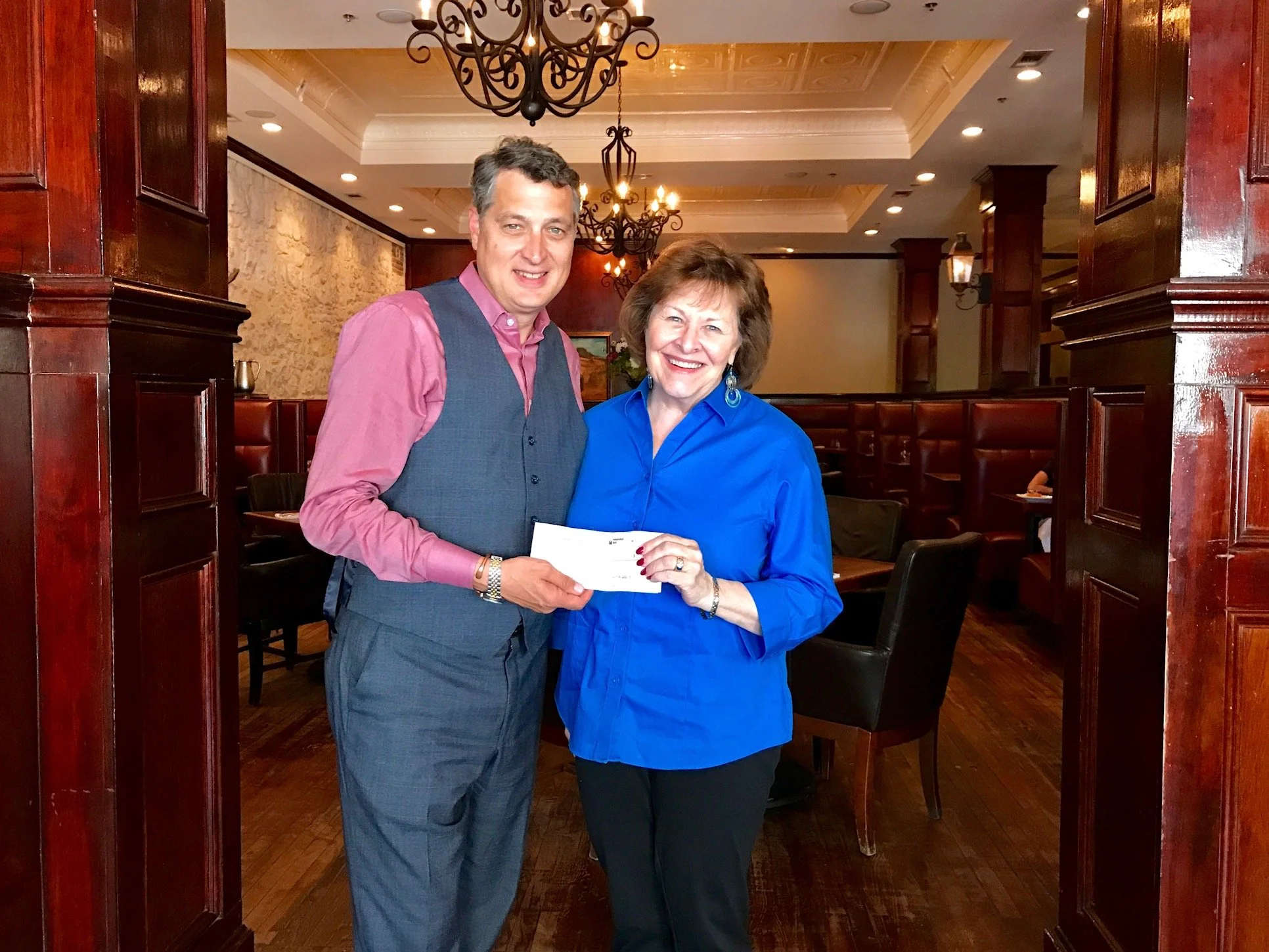 A man and a woman standing in a restaurant, smiling, holding a check, with chandeliers and wooden decor in the background.