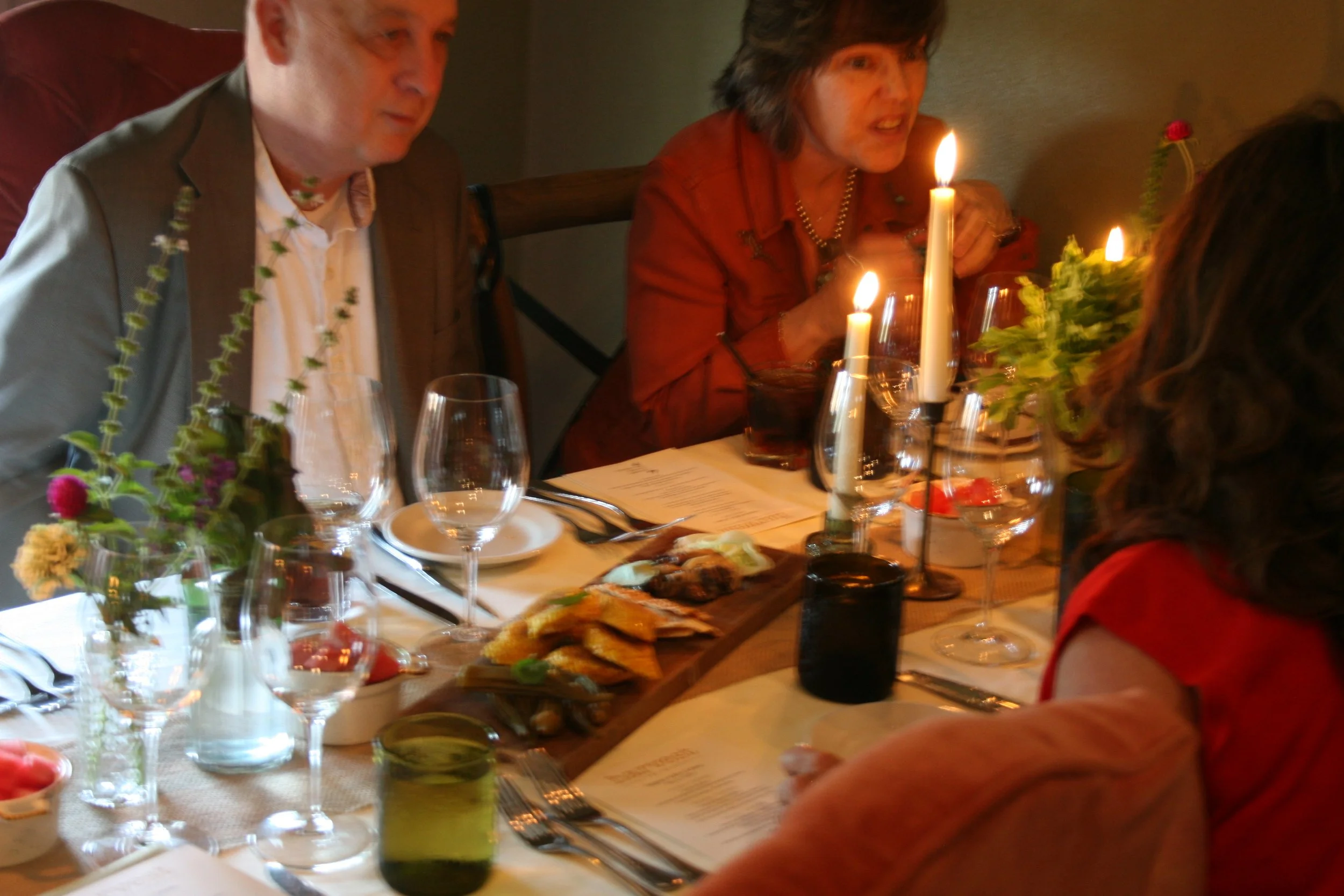 People sitting at a dining table with lit candles, flowers, wine glasses, and a variety of foods, engaged in conversation.