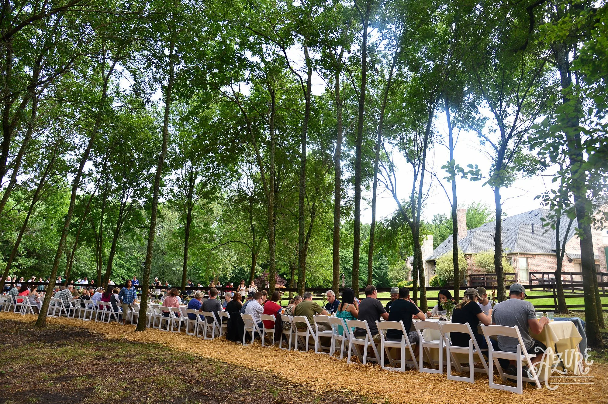 Outdoor gathering of people dining at long tables with white chairs in a wooded area near a house.