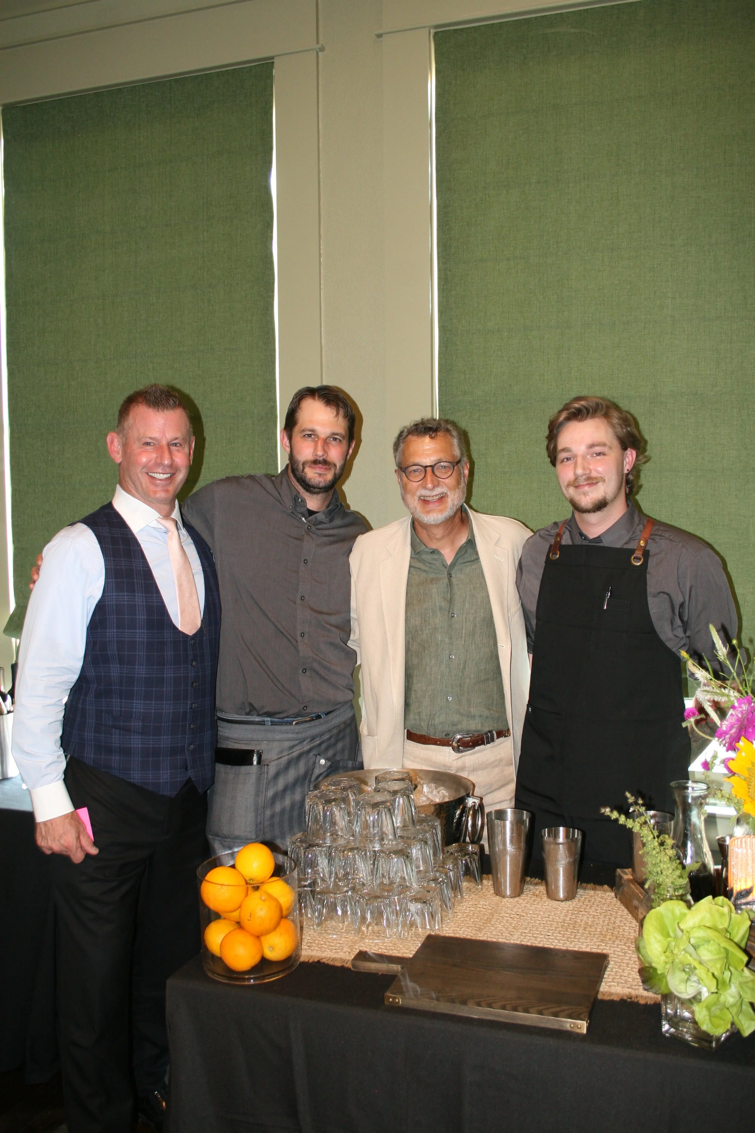 Four men smiling and standing together behind a table with glasses, oranges, and flowers at an indoor event.