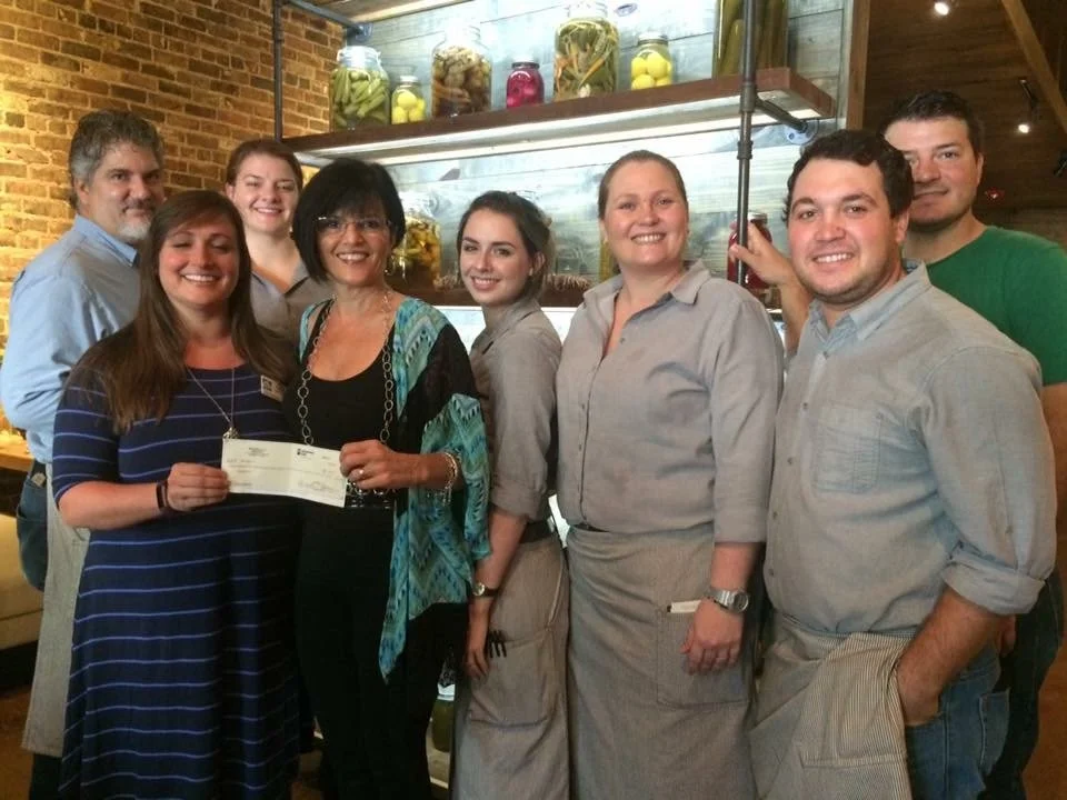 Group of eight diverse people standing together in a restaurant, smiling at the camera, with a brick wall and jars of preserved foods on shelves in the background.