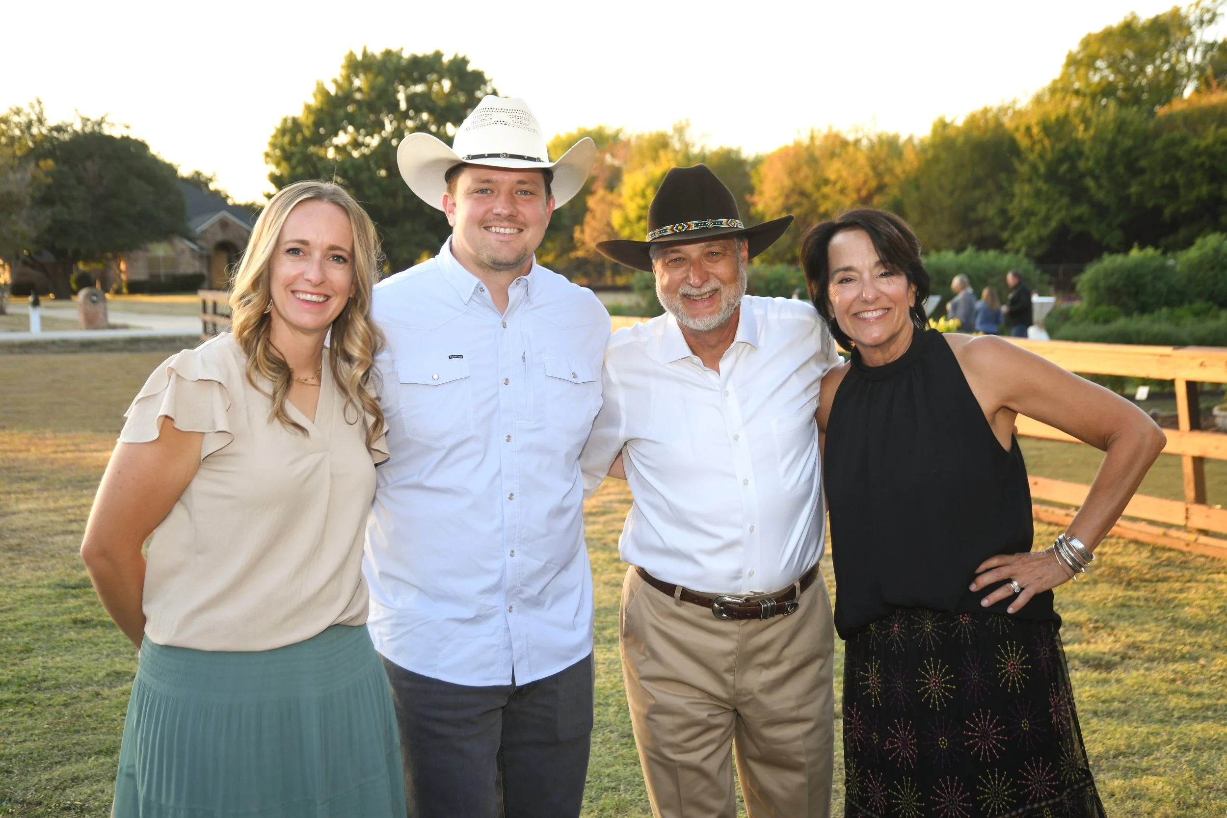 Four smiling people standing outdoors in a park, with trees and a fence in the background, during sunset. Two men and two women, dressed casually, with two wearing cowboy hats.