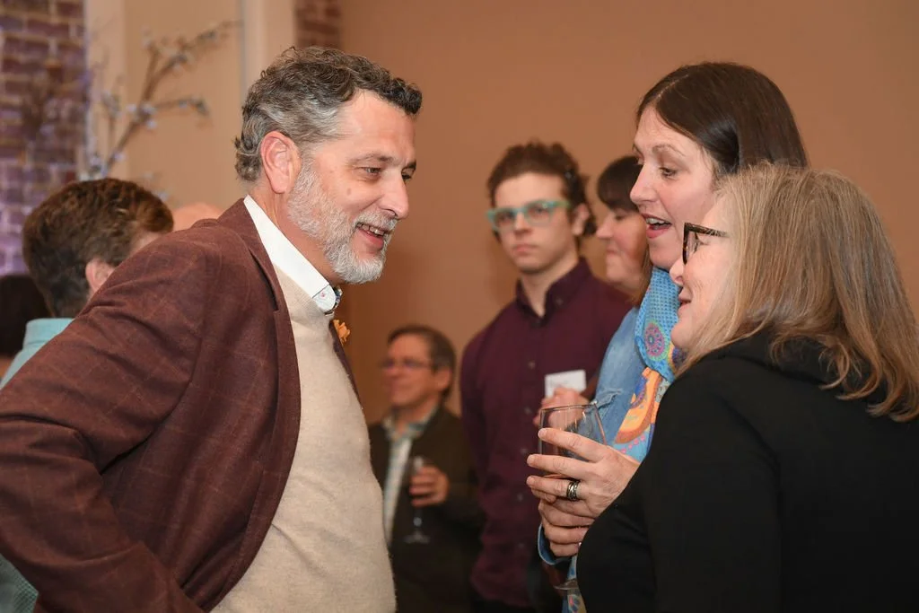 Three people engaged in conversation at a social gathering, with other attendees in the background.