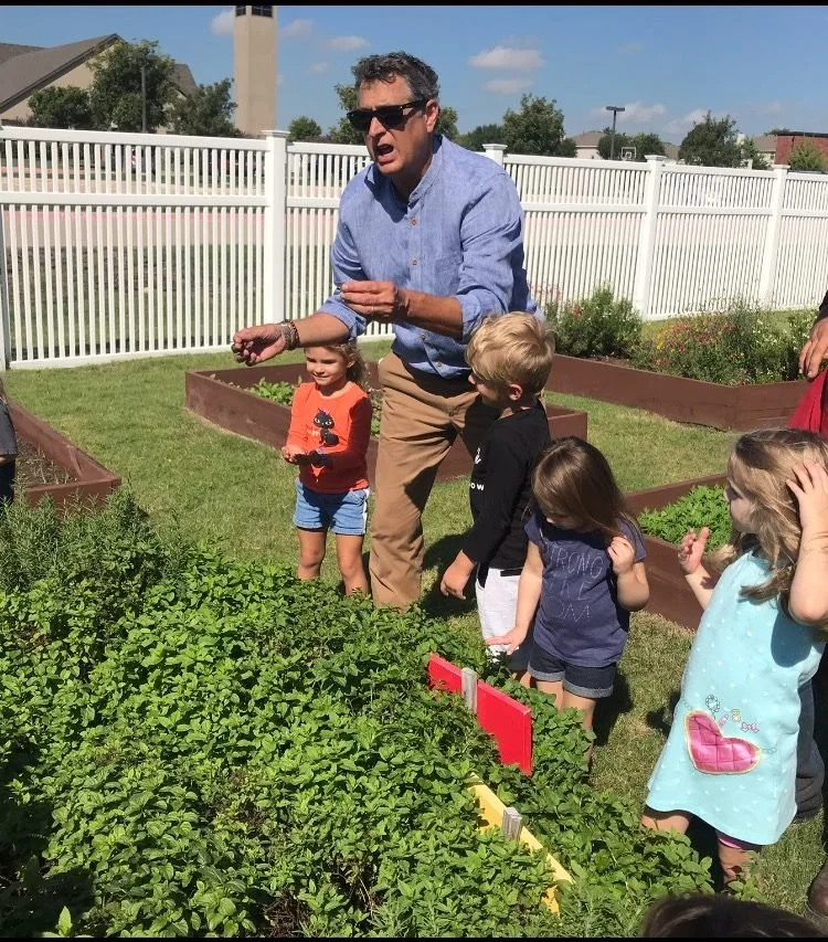 A man leading a garden activity with young children in a backyard with raised garden beds, plants, a white fence, and blue sky.